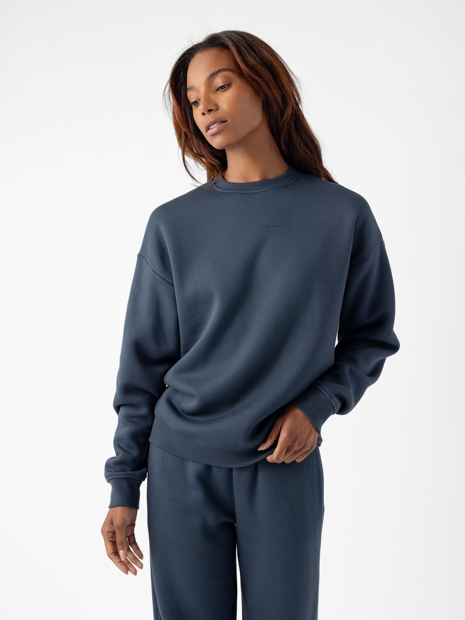 A person with long hair stands against a plain white background, dressed in Cozy Earth's dark blue Women's CityScape Crewneck sweatshirt and matching sweatpants. They have a relaxed expression with one hand resting casually by their side and the other touching the hem of the sweatshirt. 