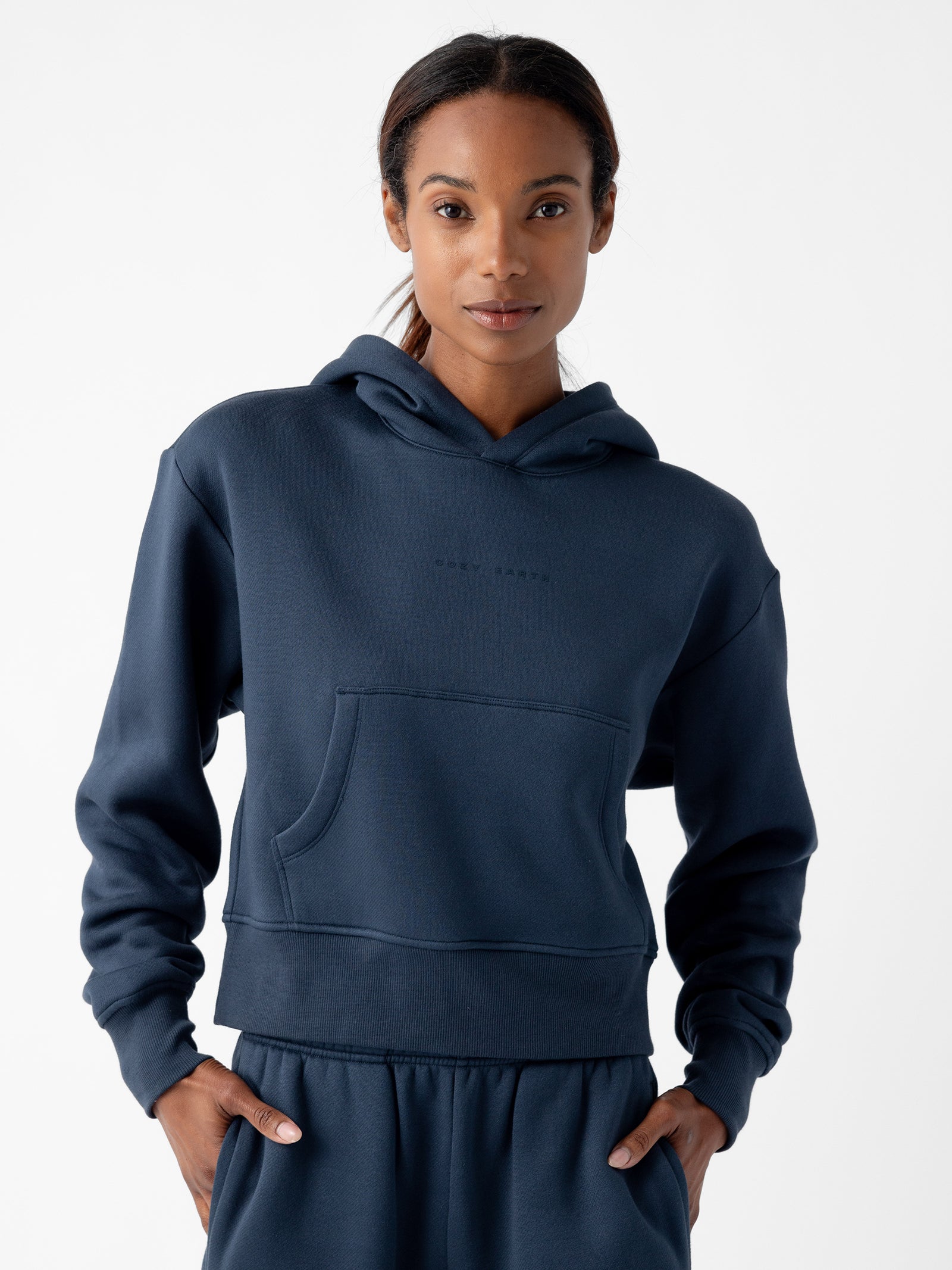 A person wearing a dark blue Women's CityScape Cropped Hoodie by Cozy Earth and matching joggers stands facing the camera with their hands in the front pocket of the hoodie. The background is white and minimalist. 