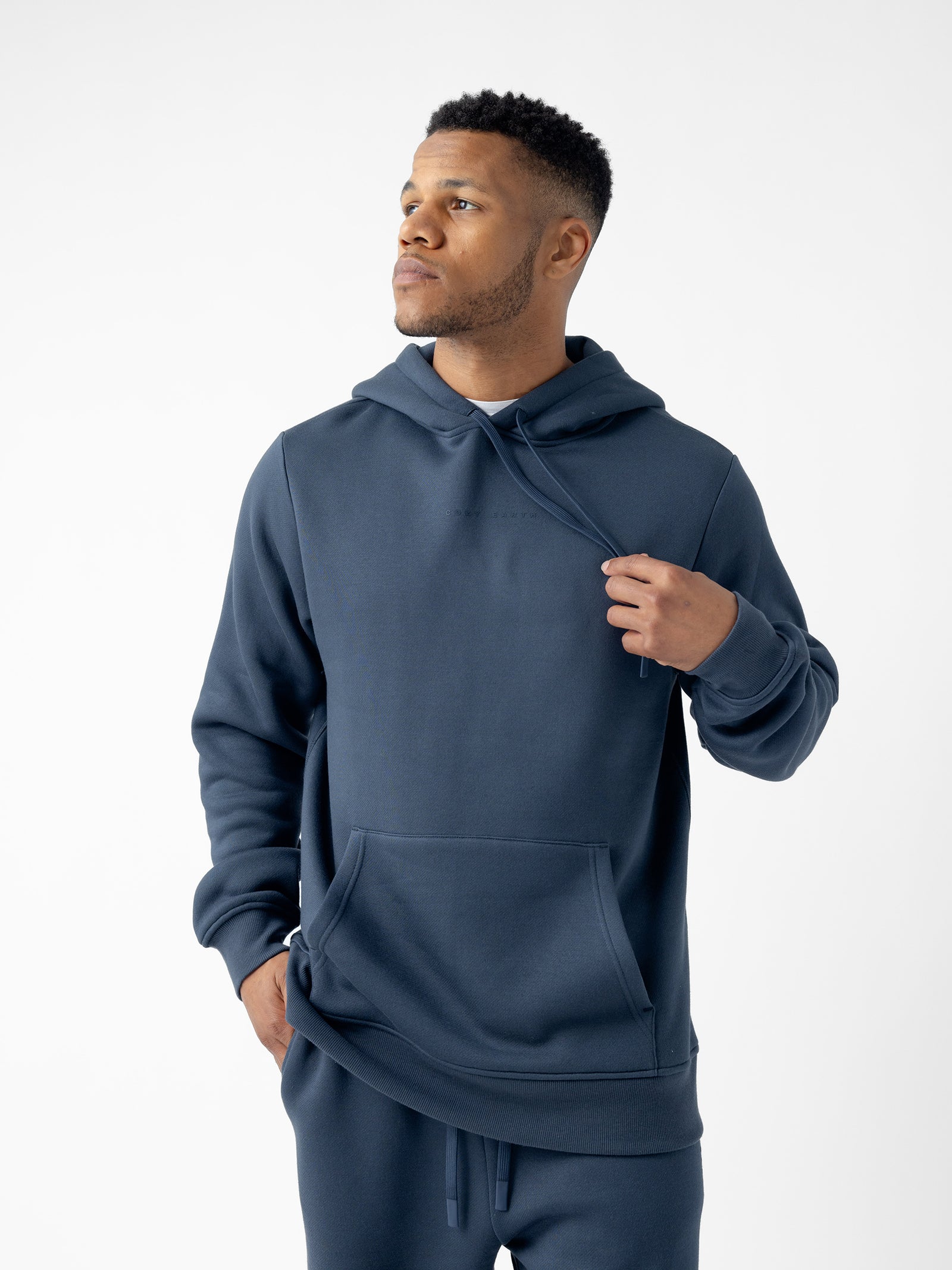 A man wearing a Cozy Earth Men's CityScape Hoodie and matching jogger pants stands against a white background. He is looking off to the side while casually holding the drawstring of his hoodie. 