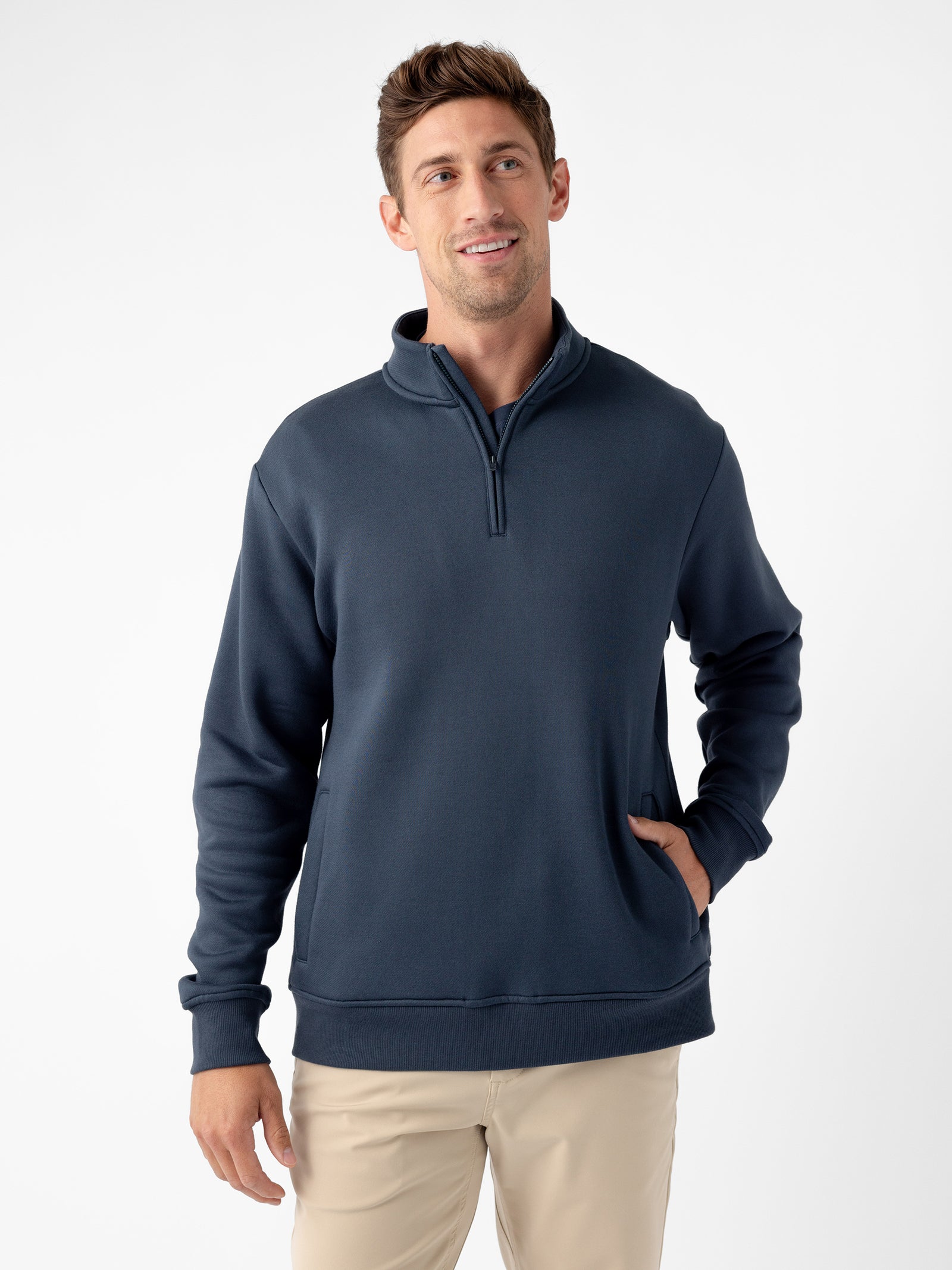 A man with short brown hair is wearing a dark blue Cozy Earth Men's CityScape Quarter Zip and light beige pants. He is standing against a plain white background with one hand in the pocket of his quarter zip and a slight smile on his face. 