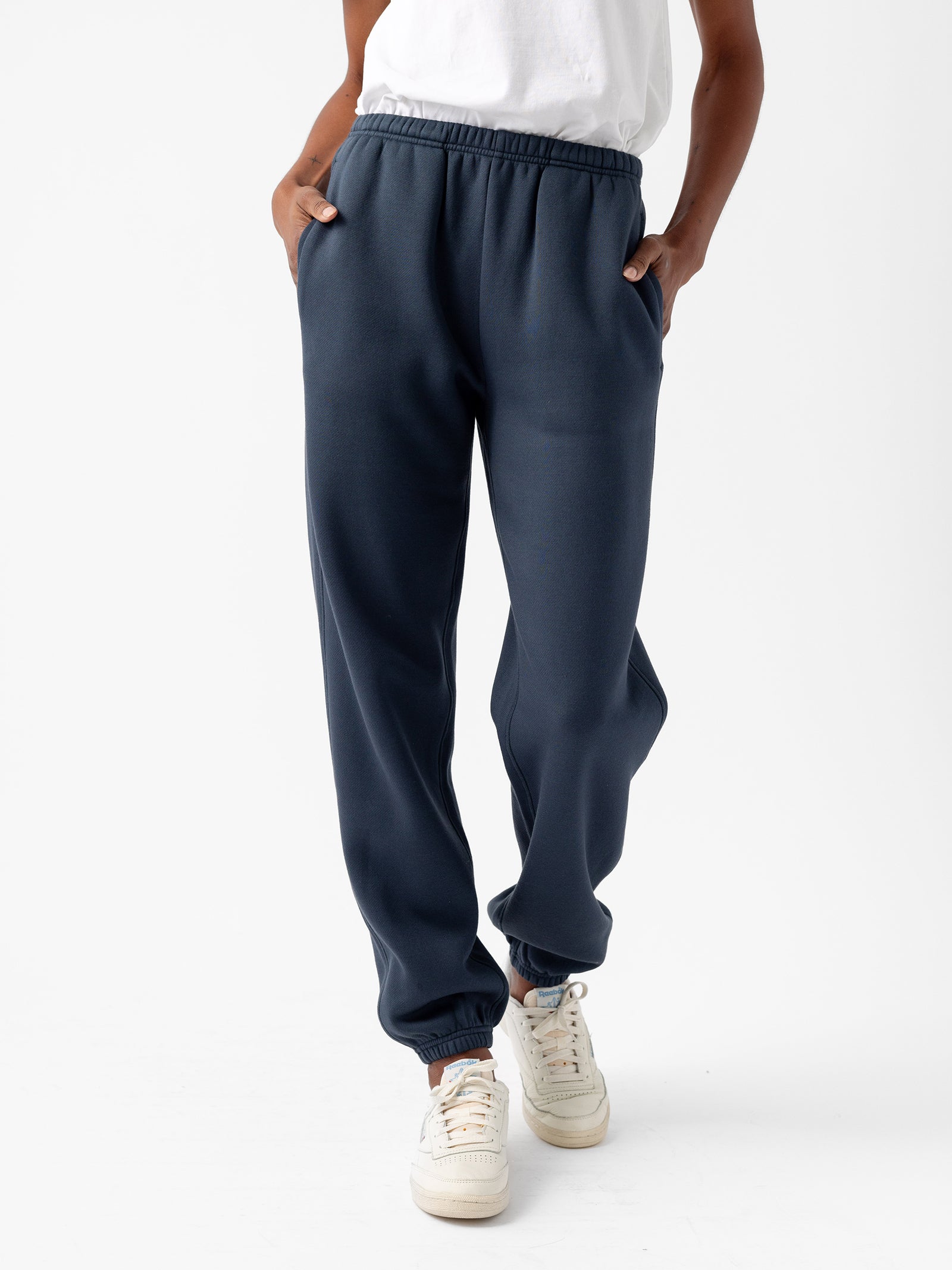 A person is wearing the Women's CityScape Sweatpant in dark blue from Cozy Earth with their hands in the pockets. They are also wearing a white T-shirt and white sneakers with colored details. The background is plain white. 