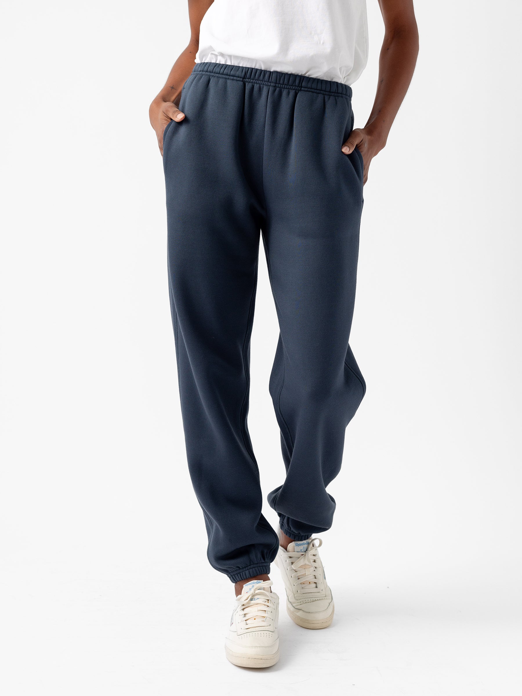 A person is wearing the Women's CityScape Sweatpant in dark blue from Cozy Earth with their hands in the pockets. They are also wearing a white T-shirt and white sneakers with colored details. The background is plain white. |Color:Eclipse