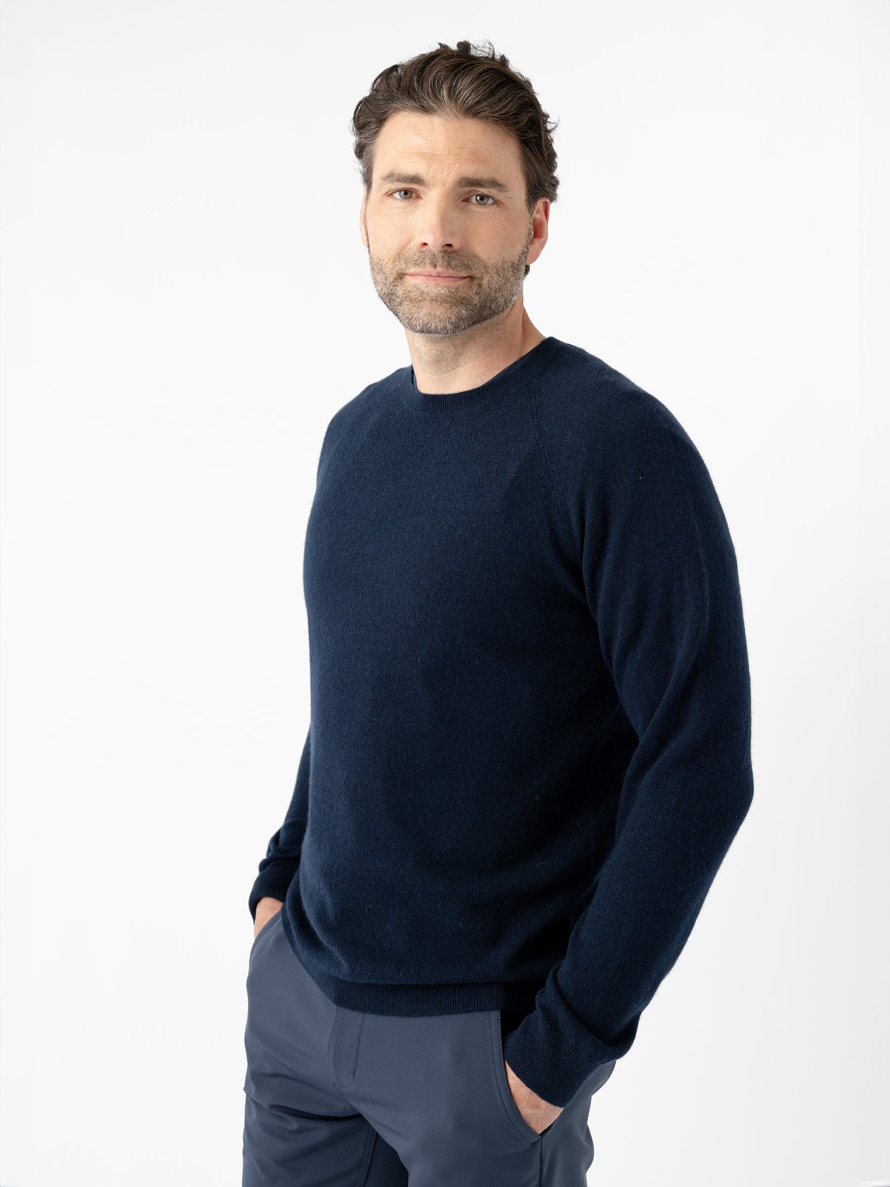 A man with dark hair and a beard stands casually with his hands in his pockets. He is wearing the Cozy Earth Men's Crewneck Sweater in navy blue, paired with gray pants. He looks at the camera with a neutral expression, set against a plain white background. |Color:Eclipse
