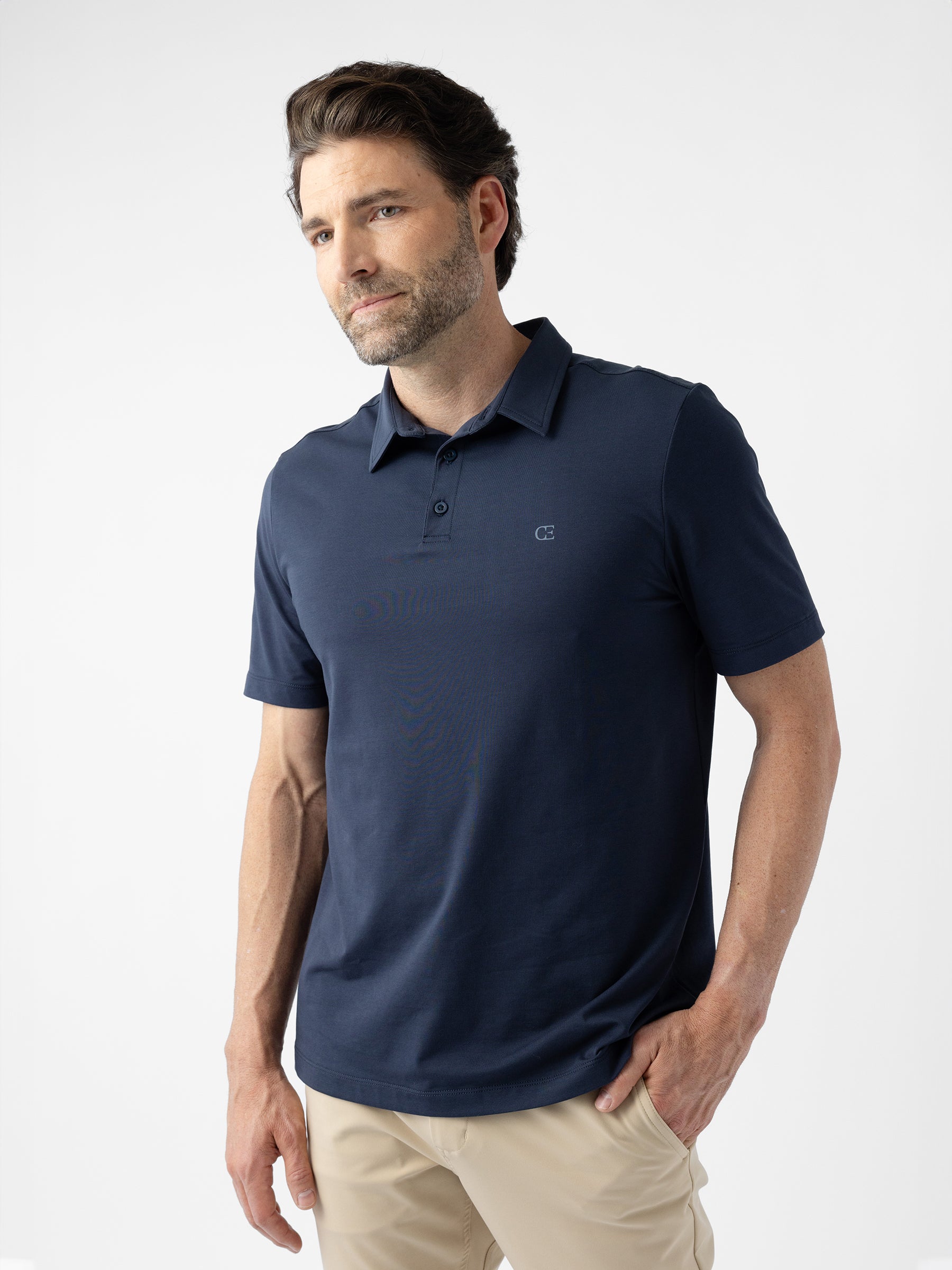 A man with a beard is wearing Cozy Earth's Men's Everyday Polo in dark navy and light beige pants. He is standing against a plain white background with one hand in his pocket, looking slightly to his left. |Color:Eclipse