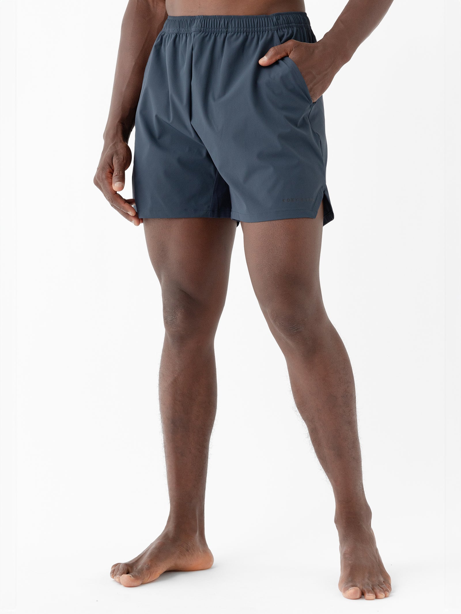 A person wearing Cozy Earth's Men's Performance Sleep Short in dark blue with hands in pockets, viewed from the waist down. The shorts have an elastic waistband and a relaxed fit. The person is barefoot and standing against a plain white background. 