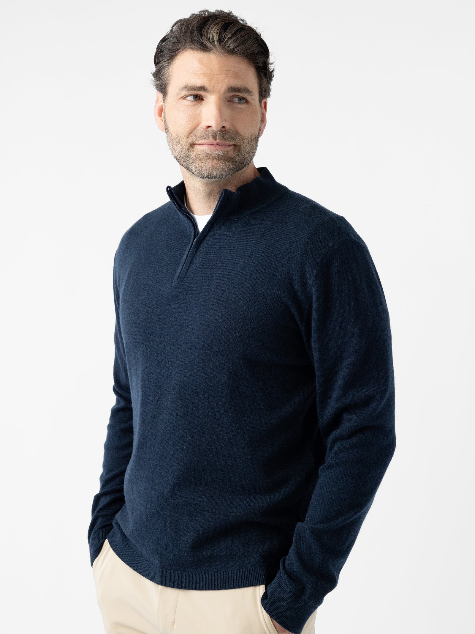 A man with dark hair and a short beard is wearing a navy blue Men's Quarter Zip Sweater from Cozy Earth paired with beige pants. He stands with his hands in his pockets against a plain white background, looking to his right with a slight smile. 