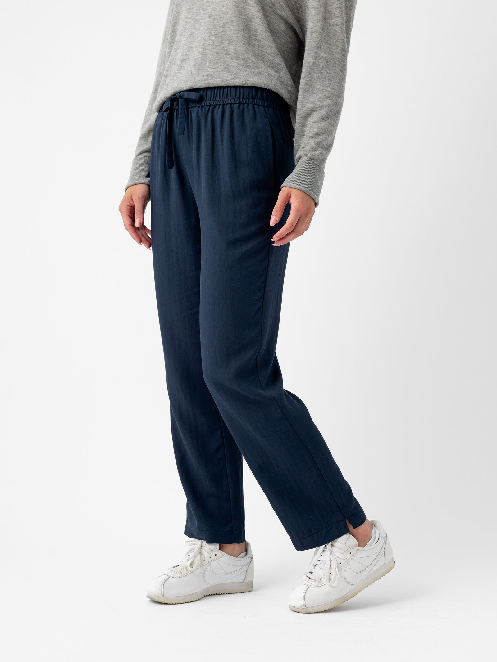 A person wearing a gray long-sleeve top and the Women's Sunset Cropped Pant from Cozy Earth in navy blue stands against a white background. They are also wearing white sneakers. The person's face is not visible. 