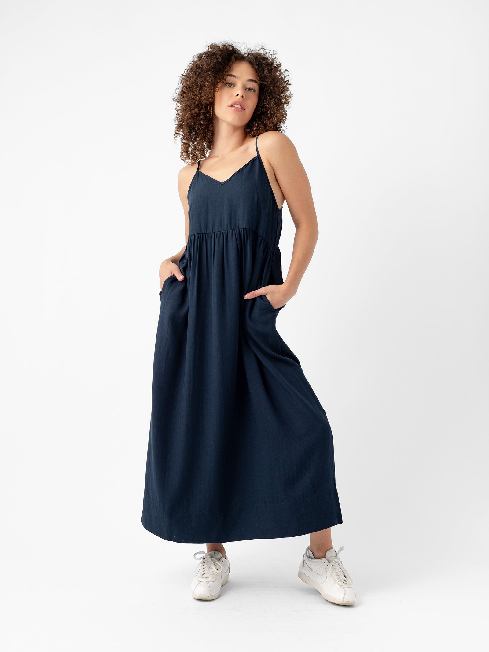 A person with curly hair is standing in a confident pose, wearing the Women's Sunset Gathered Dress by Cozy Earth, which is dark blue, sleeveless, and ankle-length with thin straps. They have their hands in the dress pockets and are sporting white sneakers. The background is plain white. 