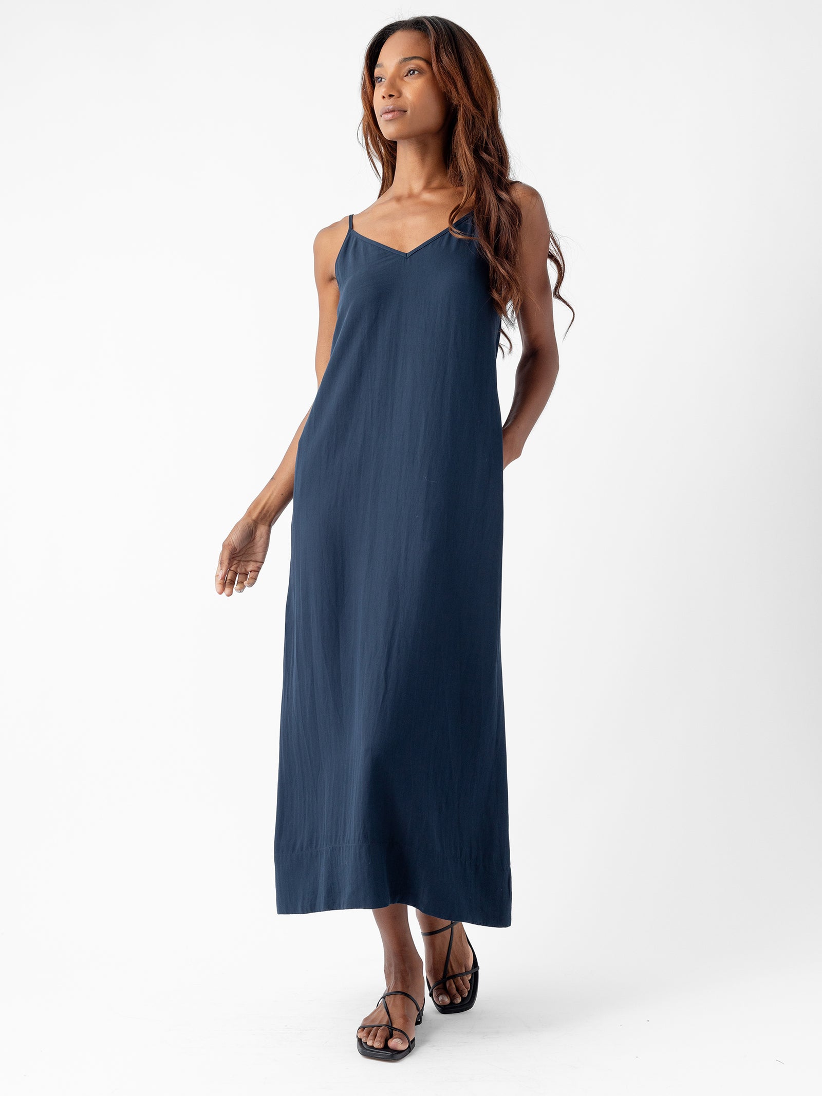 A person with long, wavy hair is wearing a Cozy Earth Women's Sunset Slip Dress in navy blue and black sandals. They are standing against a white background, looking confidently to the side with one hand in their pocket and the other arm relaxed by their side. 