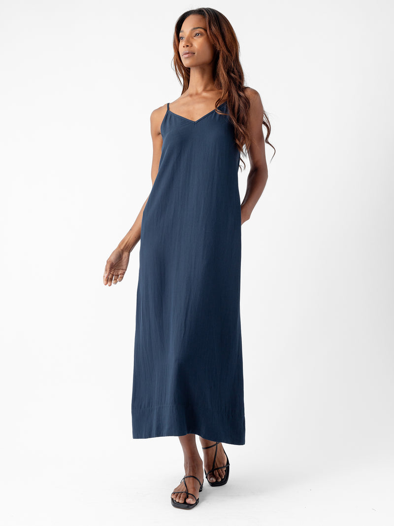 A person with long, wavy hair is wearing a Cozy Earth Women's Sunset Slip Dress in navy blue and black sandals. They are standing against a white background, looking confidently to the side with one hand in their pocket and the other arm relaxed by their side.