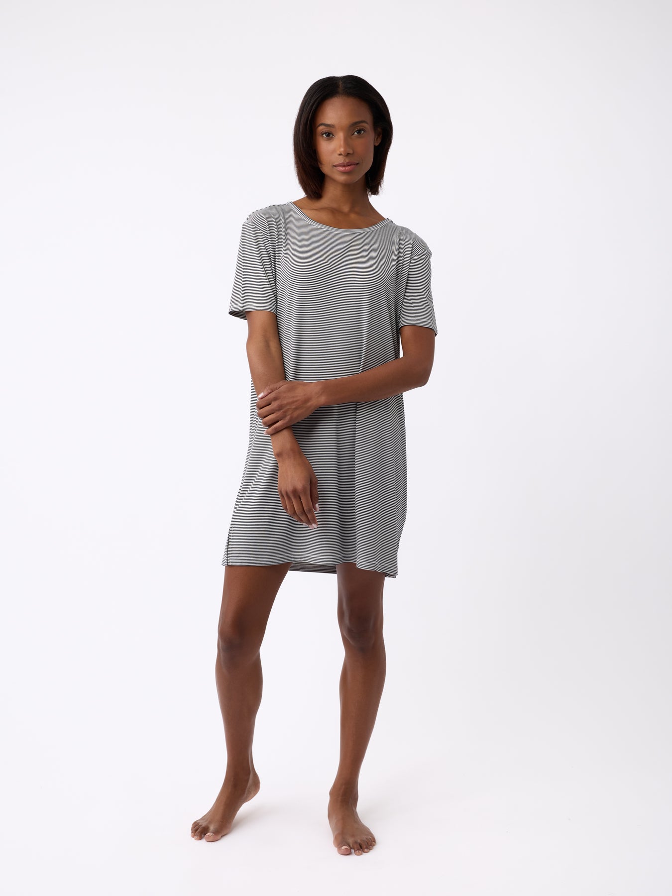 A person wears a Cozy Earth Women's Bamboo Stretch-Knit Sleep Dress, light gray and knee-length, standing barefoot against a plain white background with arms gently crossed. The short-sleeve dress offers a relaxed and comfortable fit. 