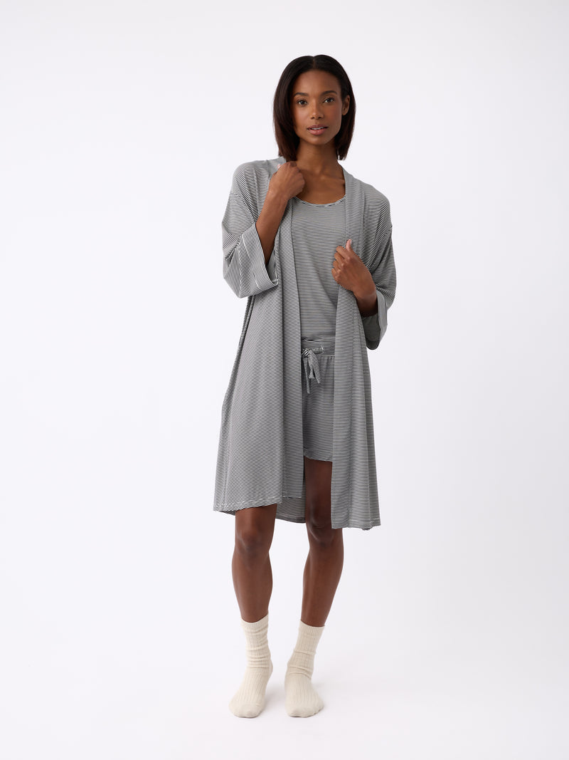 Against a plain backdrop, a person wears a Cozy Earth Women's Bamboo Stretch-Knit Kimono Robe in light gray over a matching top and shorts. They pair it with cream socks and have shoulder-length hair.