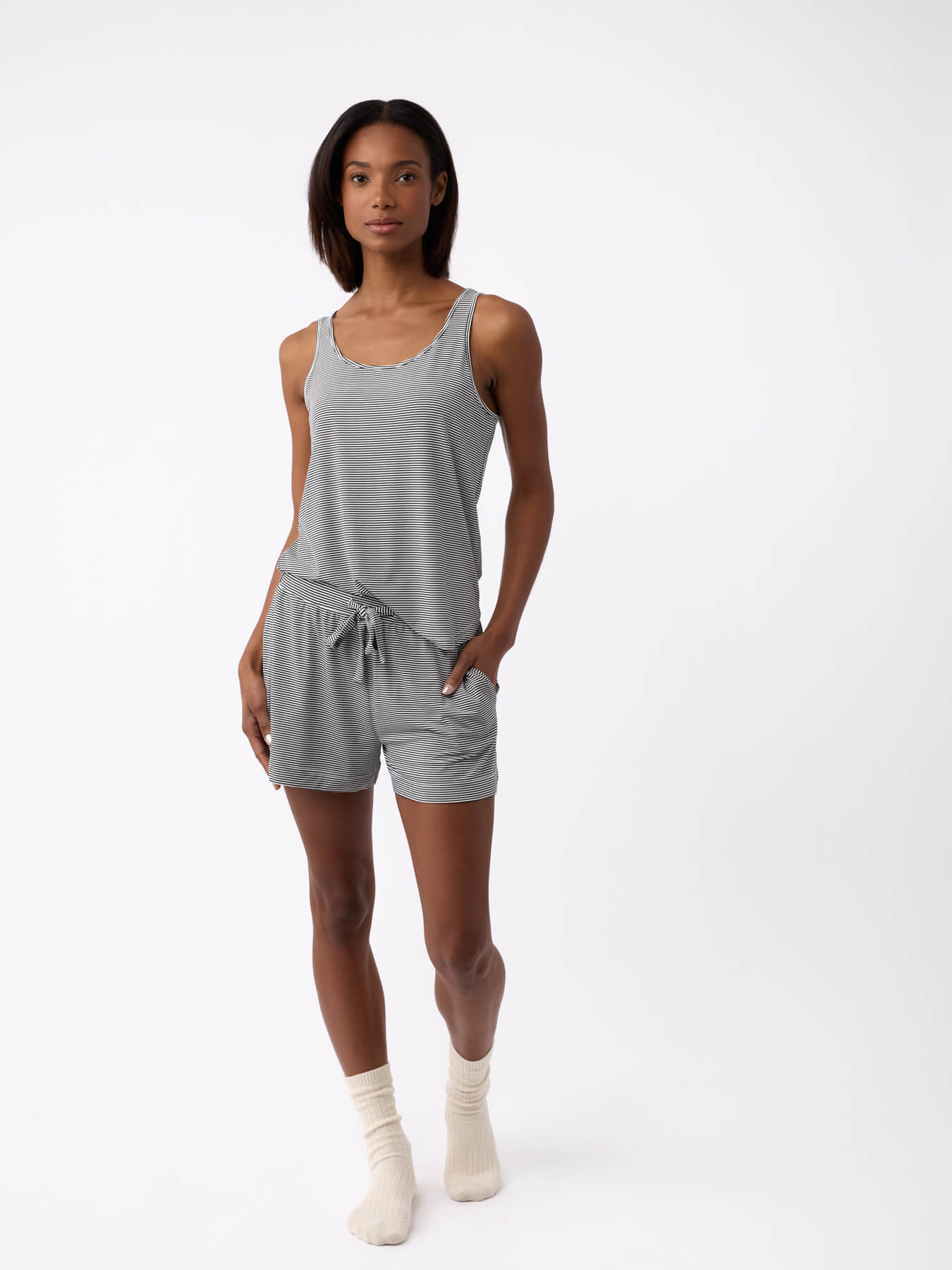 A woman stands against a plain white background wearing the Cozy Earth Women's Bamboo Stretch-Knit Lounge Tank and matching drawstring shorts, paired with light ankle socks. She has one hand in her pocket and a neutral expression.