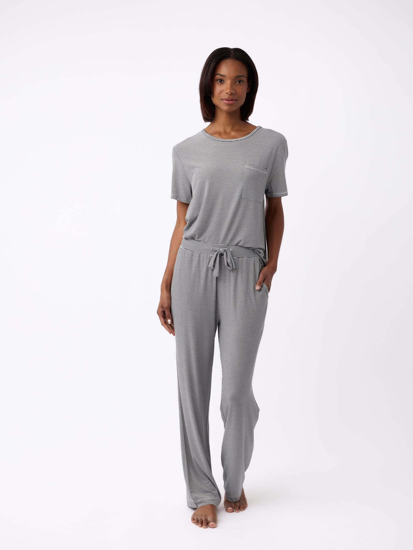 A woman in Cozy Earth's Women's Bamboo Stretch-Knit Lounge Tee and drawstring pants stands barefoot with one hand in her pocket against a plain white background. 