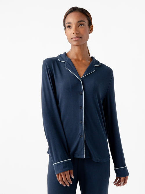 A person stands against a white background wearing the Women's Bamboo Rib Knit Classic Long Sleeve Pajama Top from Cozy Earth in navy blue, featuring light-colored piping along the collar, cuffs, and button line. The pajamas have a relaxed fit with long sleeves and a button-up top. The person has a serene expression. |Color:Eclipse