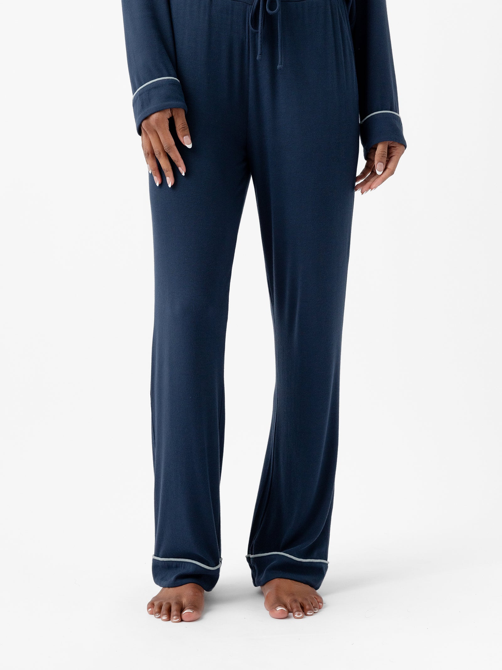 A person is standing, visible from the neck down, wearing Cozy Earth's dark blue Women's Bamboo Rib Knit Classic Pajama Pant and a matching top with white trim on the cuffs and hem. The person's hands are at their sides, and their bare feet are on a plain white background. 