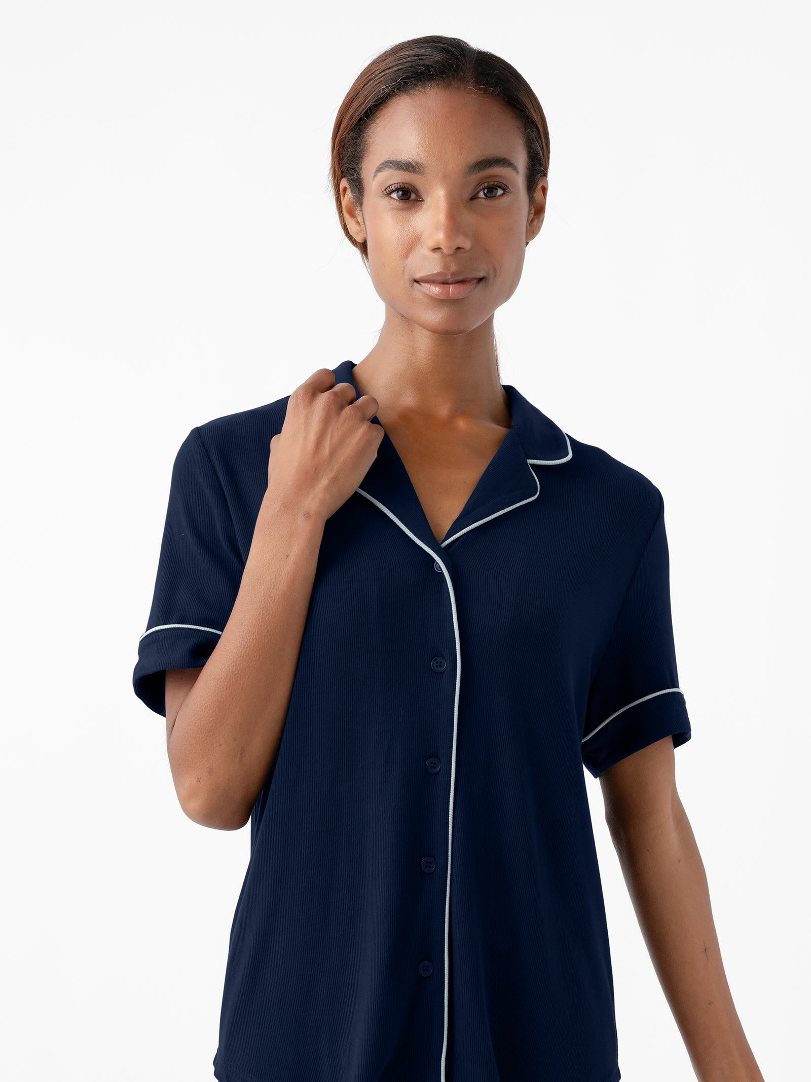 A person with a neutral expression stands against a plain white background. They are wearing Cozy Earth's Women's Bamboo Rib Knit Classic Short Sleeve Pajama Top in navy blue, featuring white piping along the collar and sleeves. One hand is gently holding the collar of the shirt. 