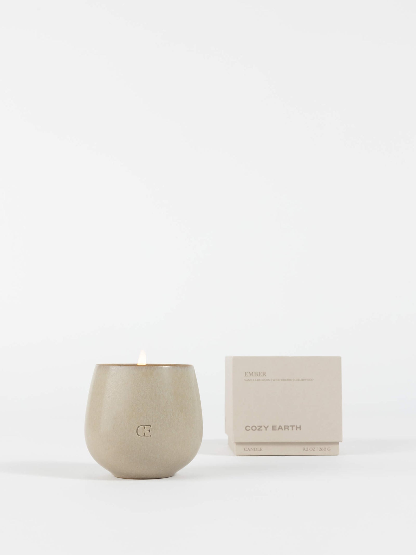 A beige ceramic Candle EMBER from Cozy Earth with a small flame is displayed beside its matching beige box labeled "Cozy Earth" on a white background.