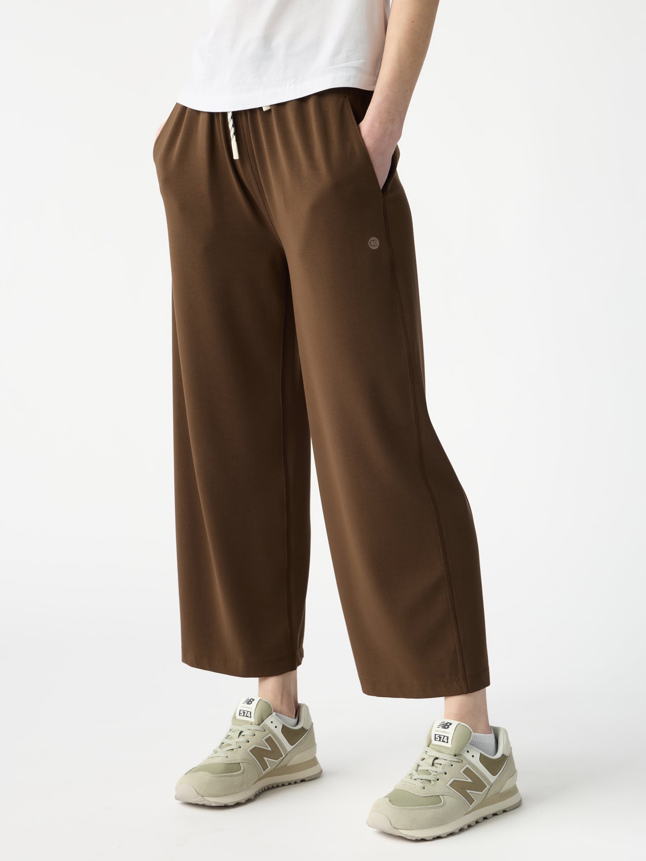 Wearing Cozy Earth's Women's Studio Cropped Wide Leg Pant in brown featuring a small circular logo on the thigh and beige New Balance sneakers, this person stands against a plain white background with hands in pockets. A glimpse of a white shirt is visible. |Color:Espresso