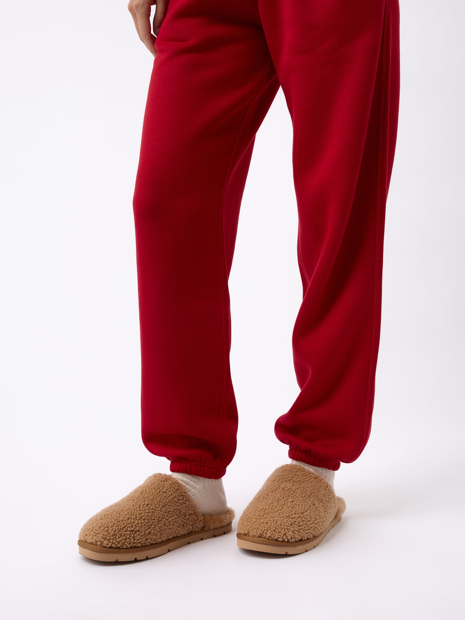 The image shows a person wearing Cozy Earth's Puffy Sheep Slippers and bright red sweatpants. The person is standing against a plain white background. The slippers have a textured, fuzzy exterior, and the sweatpants are loose-fitting with elasticized cuffs. 