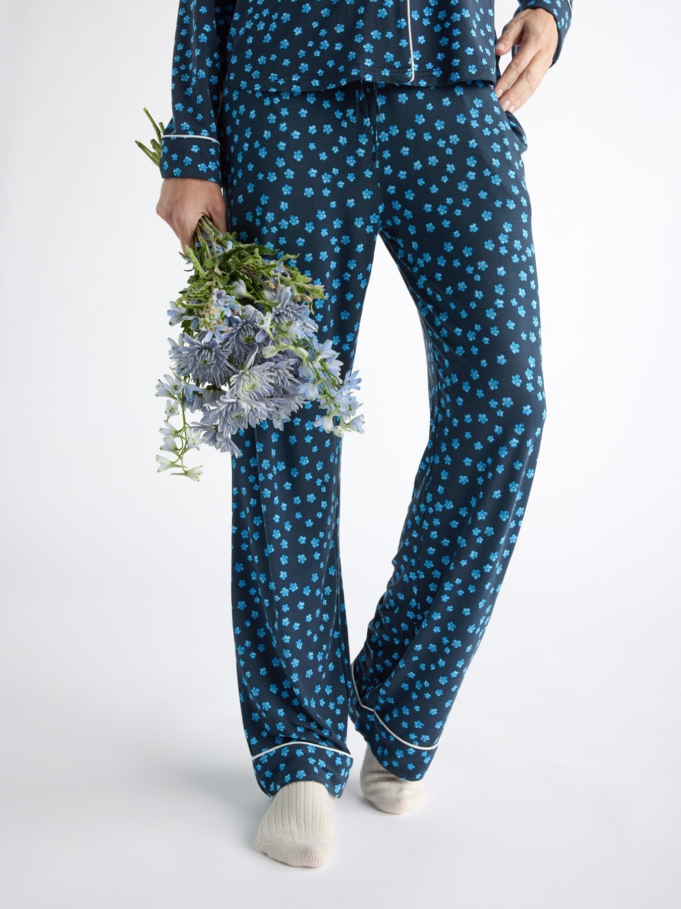 A person in Cozy Earth Women's Bamboo Stretch-Knit Pajama Pant, featuring a dark blue design with small floral patterns, holds purple flowers against a plain background while wearing light-colored socks. 