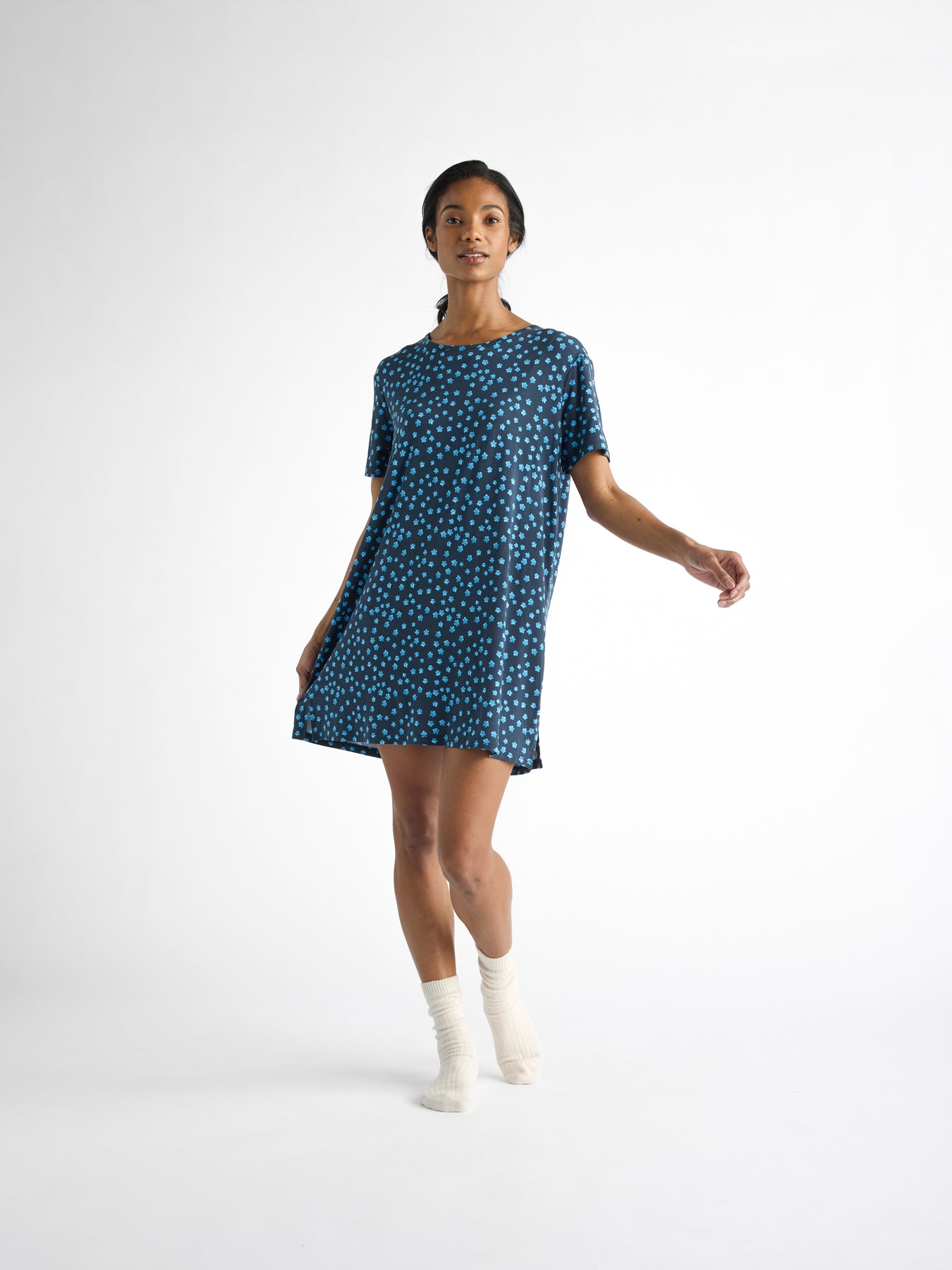 A person wears the Cozy Earth Women's Bamboo Stretch-Knit Sleep Dress in blue with white polka dots, paired with white socks, set against a plain white background. The dress, featuring short sleeves and an above-the-knee length, complements their slightly extended left arm. 