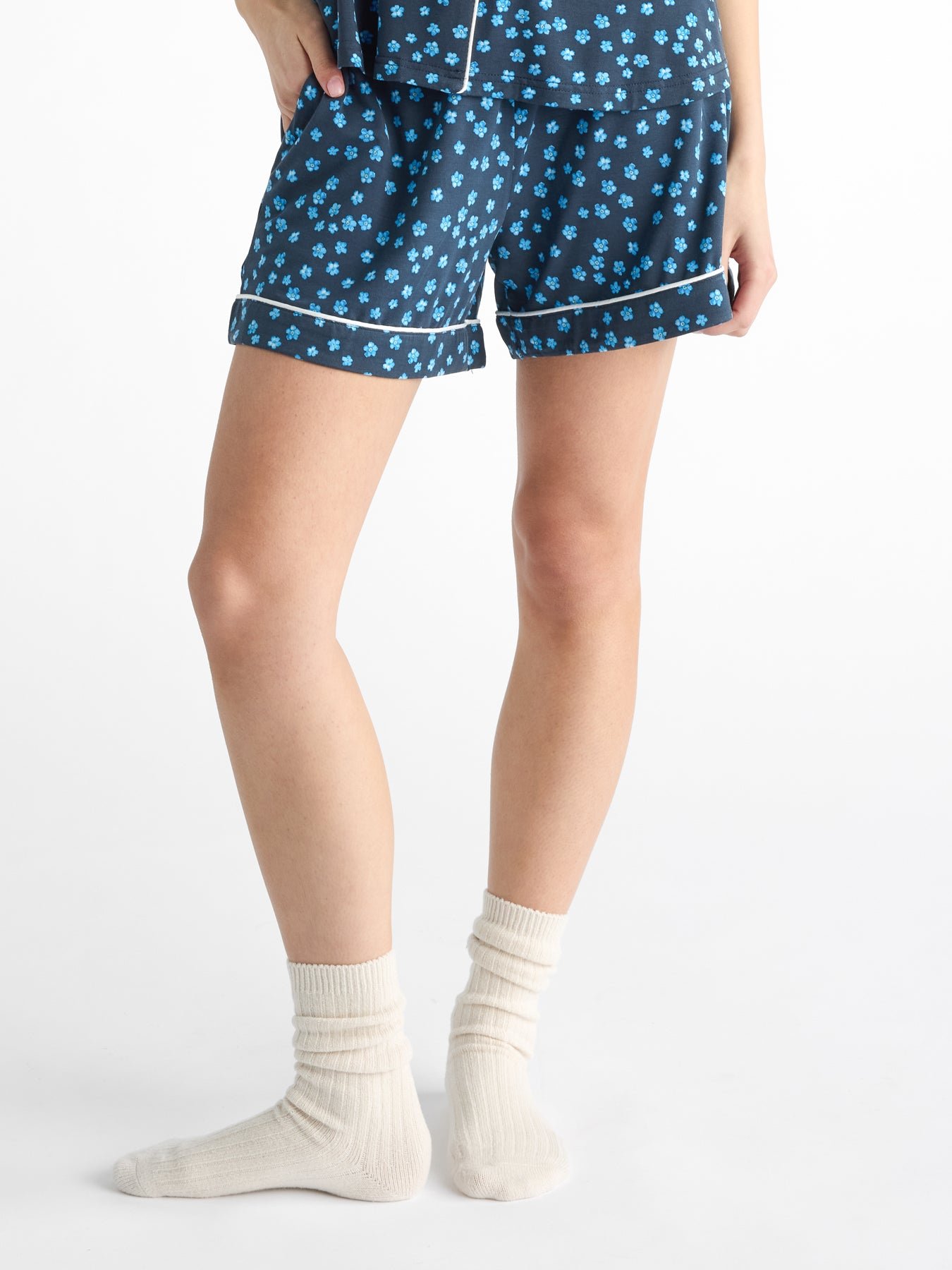 A person stands with one hand on their side, showcasing Cozy Earth Women's Bamboo Stretch-Knit Pajama Shorts in blue with a floral pattern, paired with cream socks, against a white backdrop. |Color:Forget Me Nots