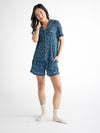 A smiling person stands against a white background, wearing a Cozy Earth Women's Bamboo Stretch-Knit Short Sleeve Pajama Set with blue polka dots and shorts, paired with white socks, while touching their chin. |Color:Forget Me Nots
