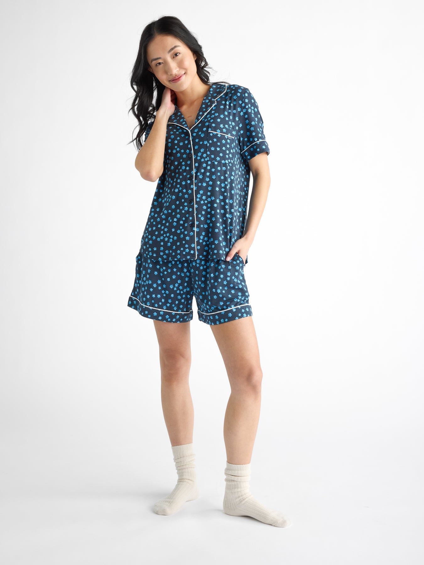 A smiling person stands against a white background, wearing a Cozy Earth Women's Bamboo Stretch-Knit Short Sleeve Pajama Set with blue polka dots and shorts, paired with white socks, while touching their chin. 