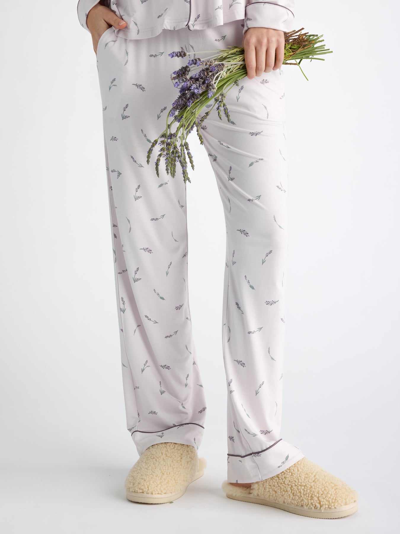 A person wearing Cozy Earth Women's Bamboo Stretch-Knit Pajama Pant with a lavender pattern and fuzzy slippers holds a small bouquet of lavender in their left hand against a plain white background. 