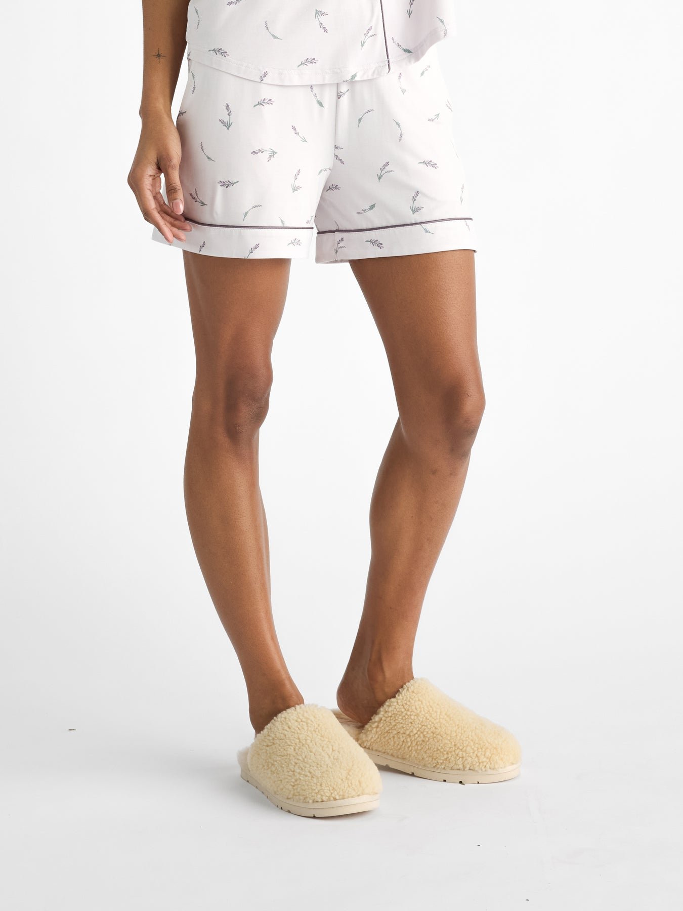 A person stands against a plain white background, wearing HIDE's Women's Bamboo Stretch-Knit Pajama Short in white with a subtle pattern and cream-colored fluffy slippers, only showing the lower half of their body. |Color:French Lavender