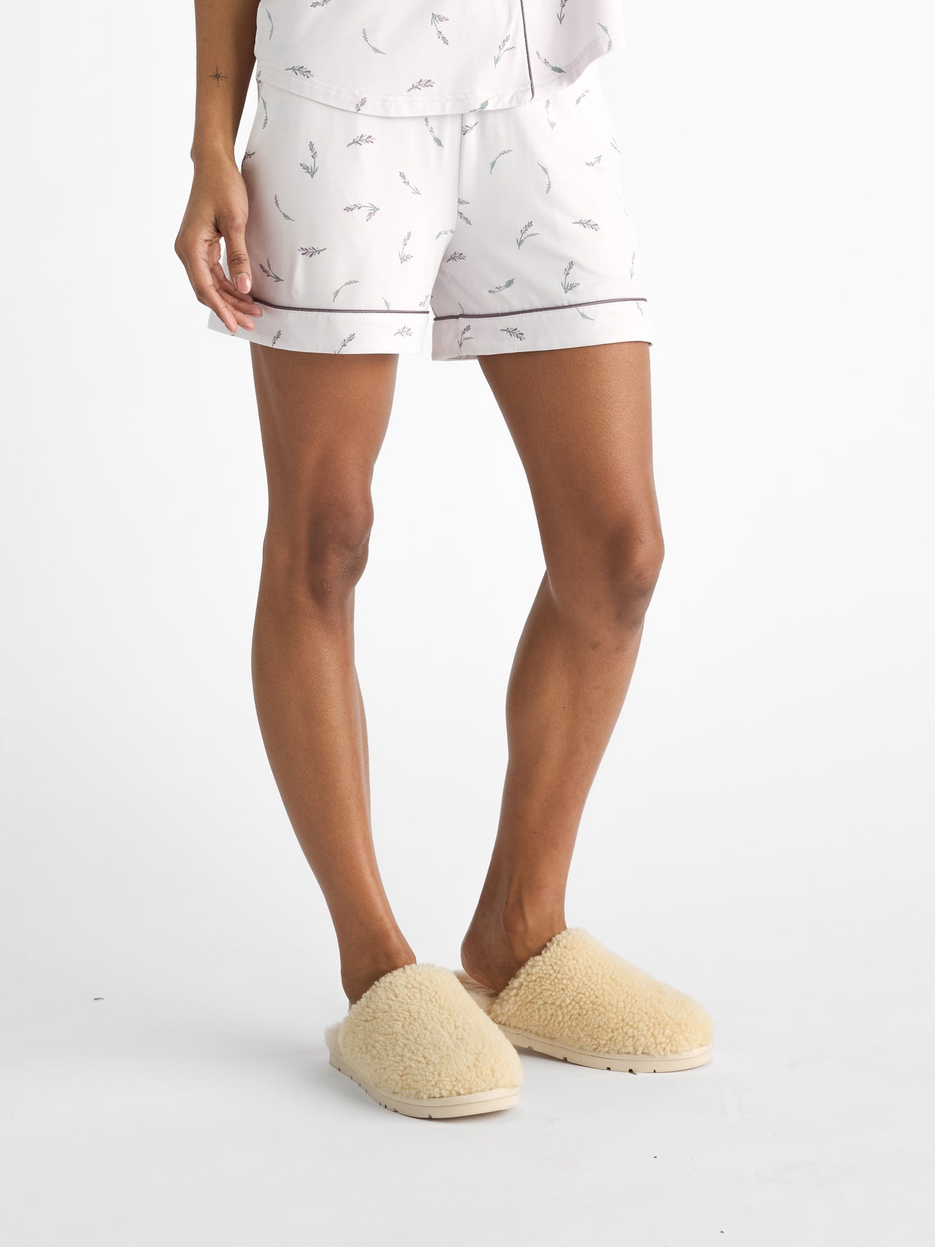 A person stands against a plain white background, wearing HIDE's Women's Bamboo Stretch-Knit Pajama Short in white with a subtle pattern and cream-colored fluffy slippers, only showing the lower half of their body. 