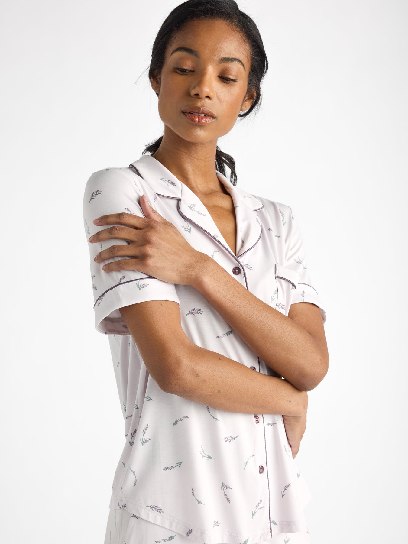 Wearing HIDE's Women's Bamboo Stretch-Knit Short Sleeve Pajama Top, featuring a subtle leaf pattern, a person stands against a plain backdrop with their arms gently hugged. Their eyes are closed, and they display a calm expression. 