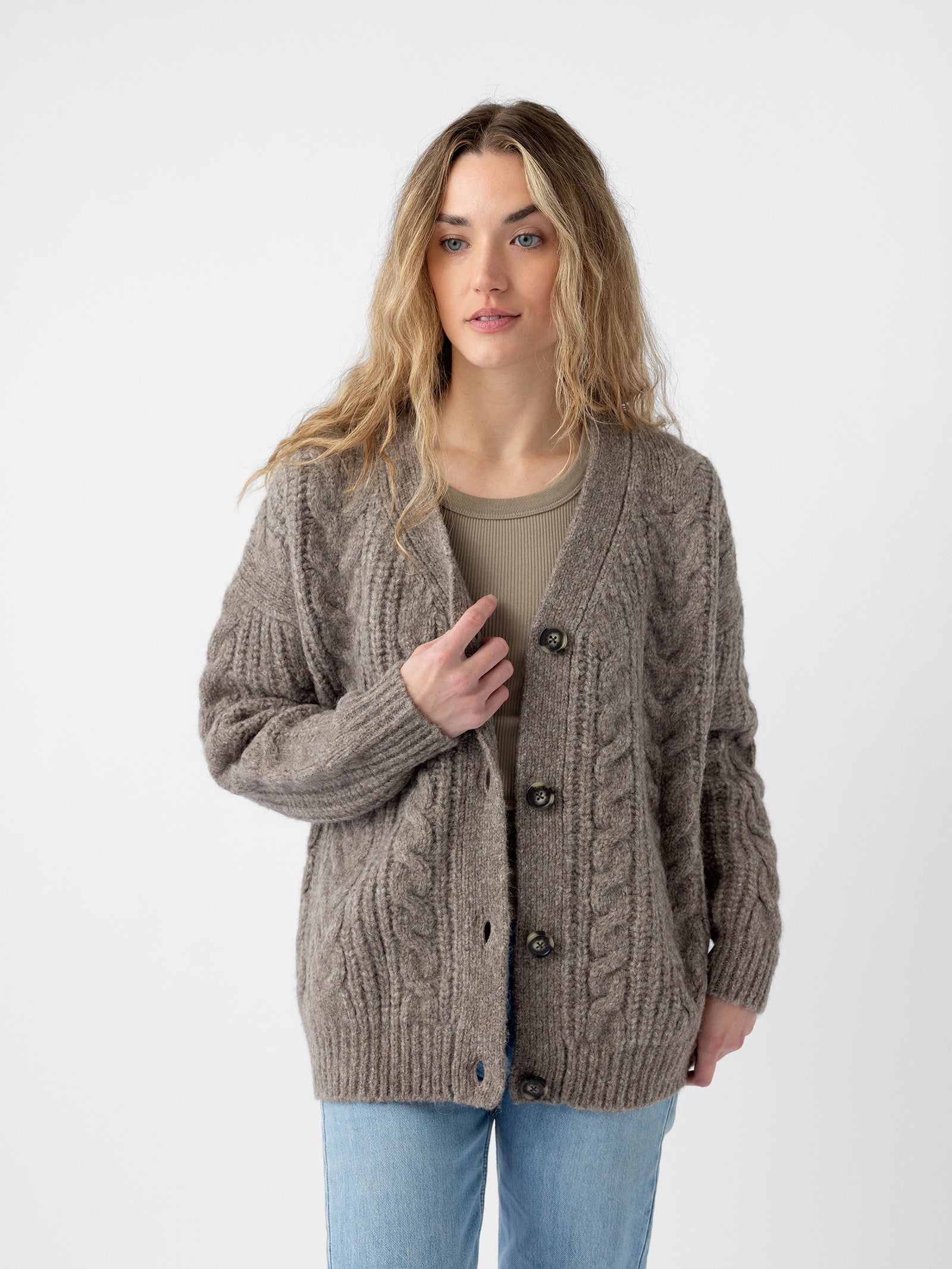 A person with long hair wearing a Cozy Earth Oversized Cable Knit Cardigan, which is thick and textured in gray-brown, over a ribbed beige top and light blue jeans stands against a plain white background. Their left hand is adjusting the cardigan. 