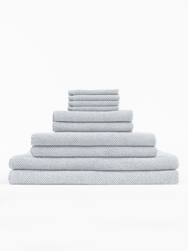 The Cozy Earth Complete Nantucket Bath Bundle features light gray textured towels in assorted sizes, neatly stacked in a pyramid formation against a plain white background.