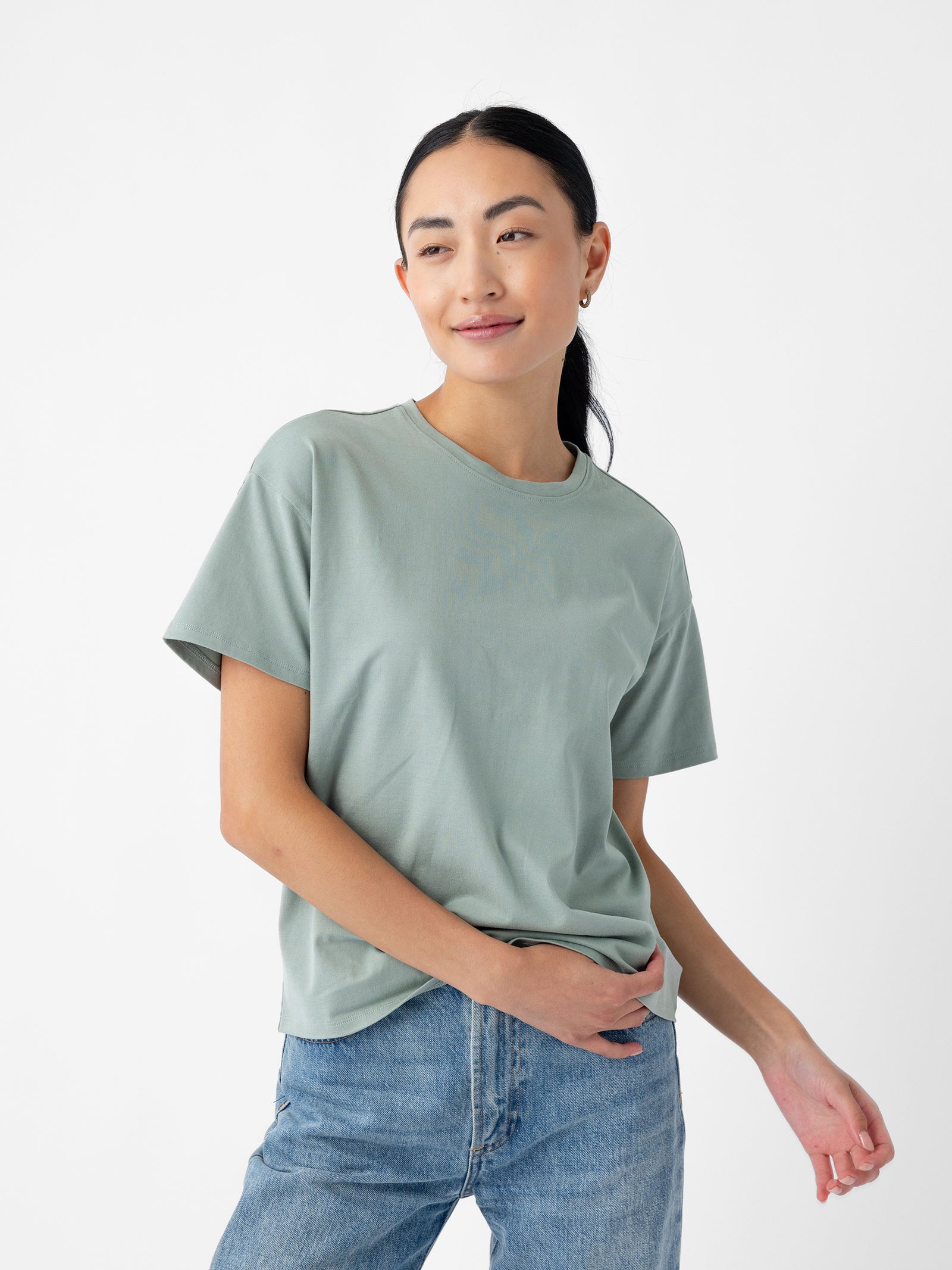 A person with long dark hair tied back is wearing the Women's All Day Tee in light green and blue jeans. They are standing against a plain white background, holding part of the t-shirt with one hand and smiling slightly. The t-shirt is by Cozy Earth. 