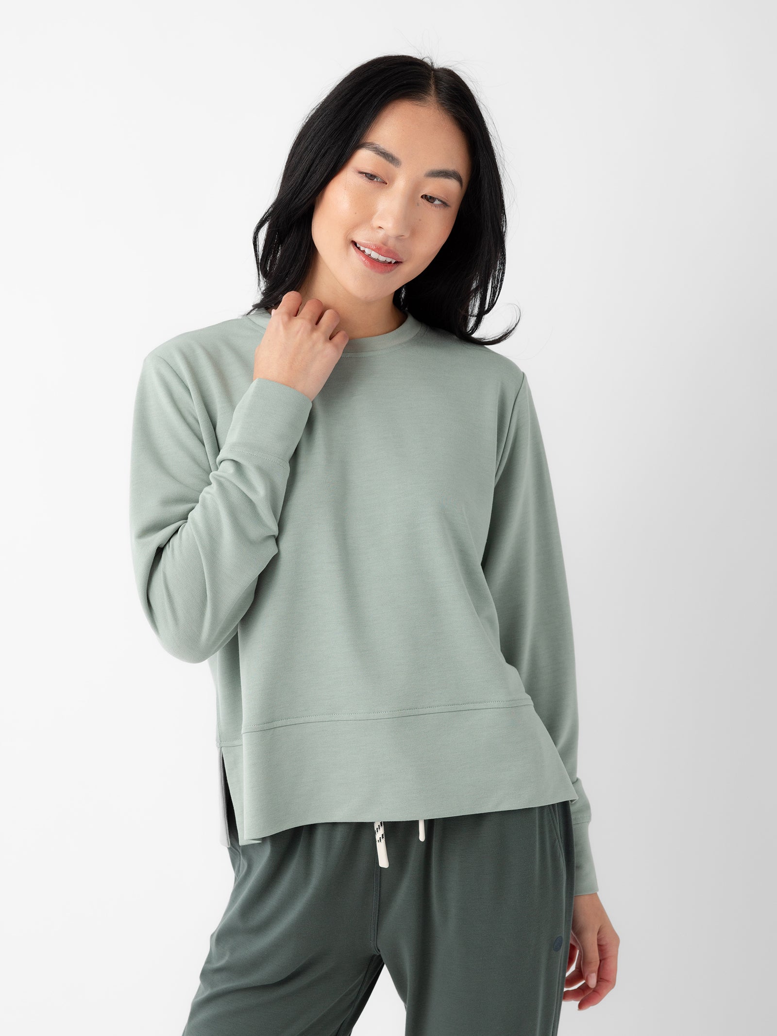 A person with long black hair is standing against a plain white background, wearing a Cozy Earth Women's StudioLite Crewneck in light green and dark green pants. They are looking down with a gentle smile, gently touching their neck with one hand. 