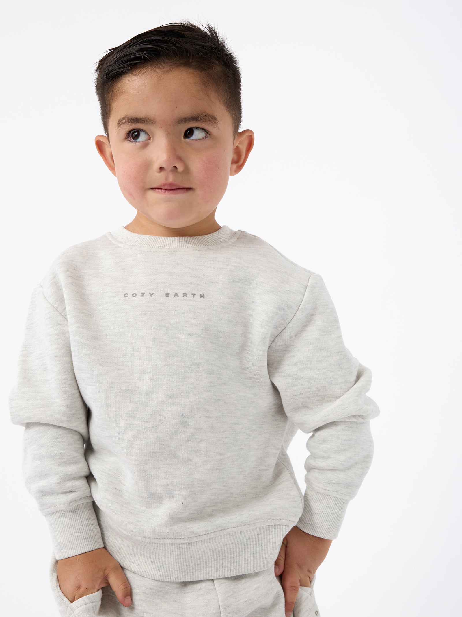 A young boy with short dark hair is wearing a light gray Kid's CityScape Crewneck by Cozy Earth. He is looking to the side with a slight smile against a plain white background. 