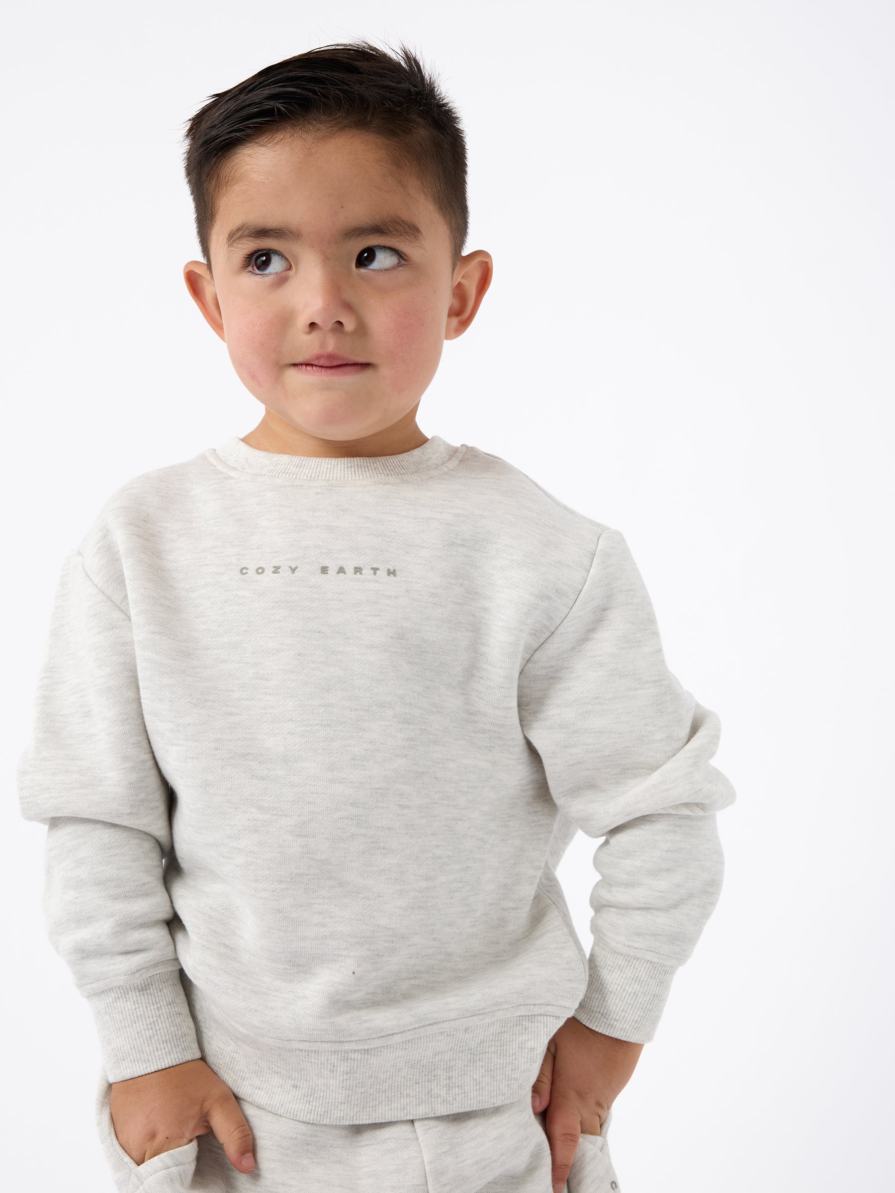 A young boy with short dark hair is wearing a light gray Kid's CityScape Crewneck by Cozy Earth. He is looking to the side with a slight smile against a plain white background. |Color:Heather Grey