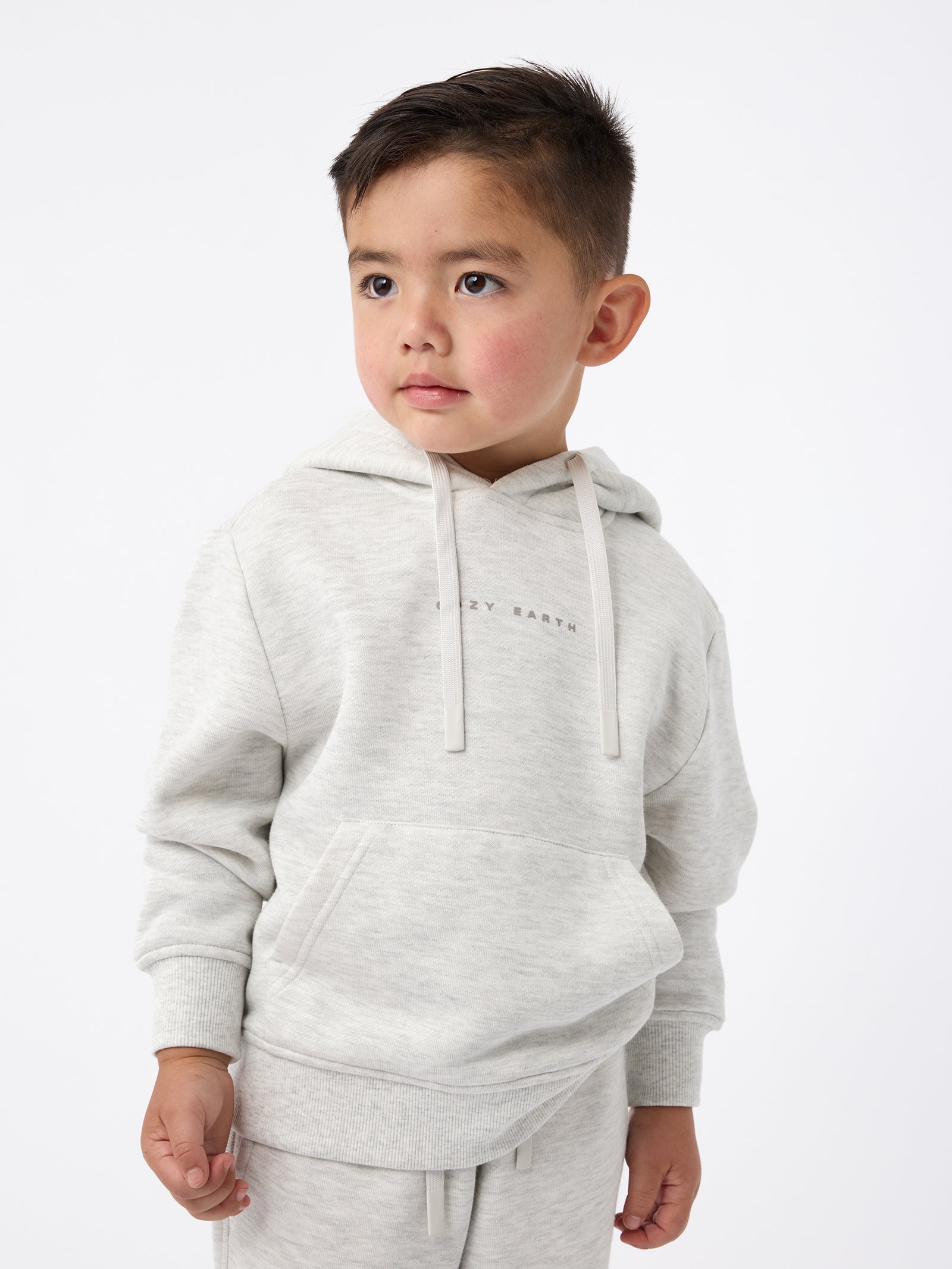 A young boy with short brown hair stands against a white background, wearing the Cozy Earth Kid's CityScape Hoodie, a light gray piece with subtle text on it, paired with matching sweatpants. He gazes to the side with a thoughtful expression. 