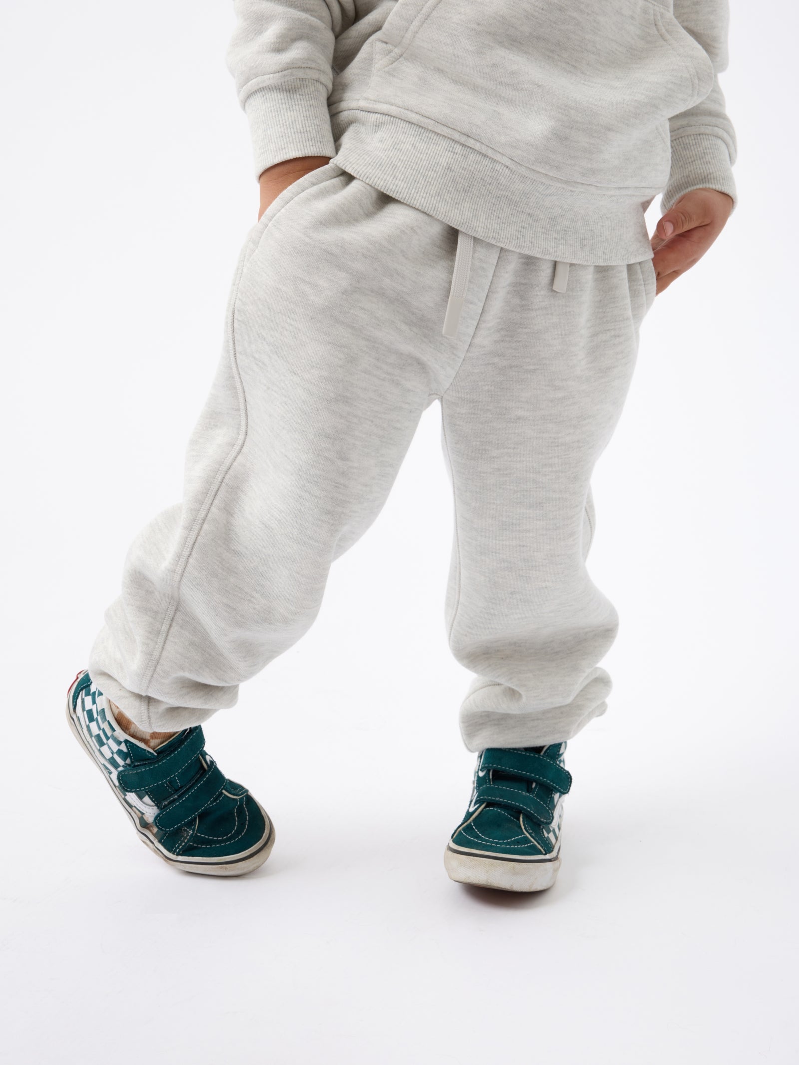 A child dressed in Cozy Earth's Kid's CityScape Sweatpant, with their hands in the pockets and sporting teal sneakers with white soles, stands against a plain white background. 