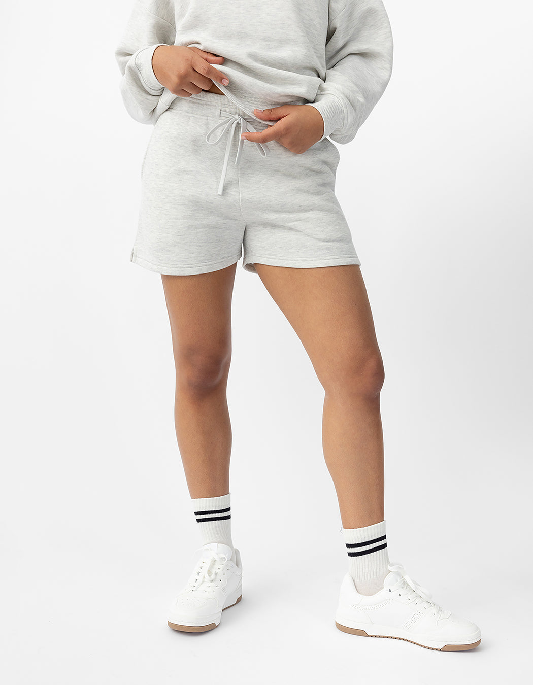 A person is wearing Cozy Earth's Women's CityScape Shorts paired with a light gray sweatshirt, white sneakers, and socks featuring black stripes. They stand against a plain white background with one hand pointing downwards. |Color:Heather Grey