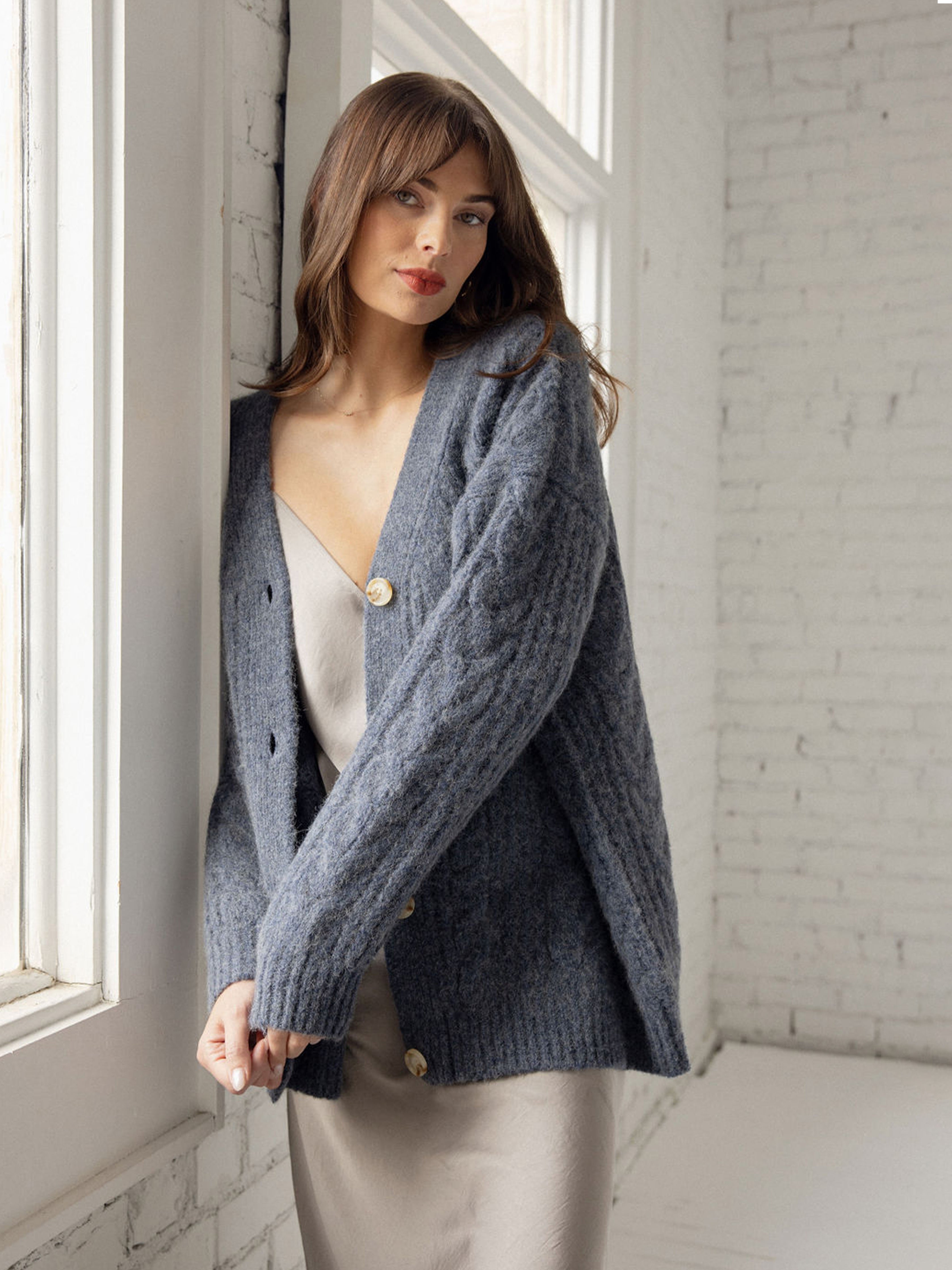 Woman wearing indigo cardigan over dress leaning against white brick wall 