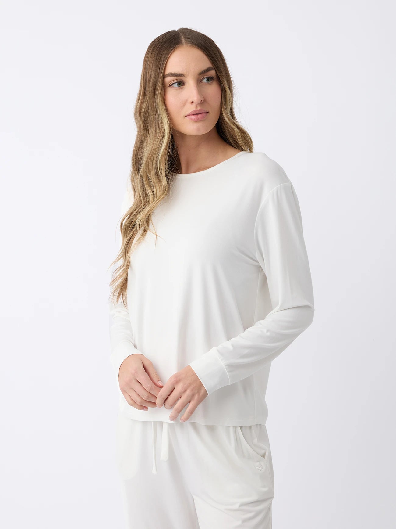 A woman with long hair, wearing a Cozy Earth Women's Stretch Knit Bamboo Long Sleeve Lounge Tee and white pants, stands against a plain background. She clasps her hands in front of her and gazes to the side. |Color:Ivory