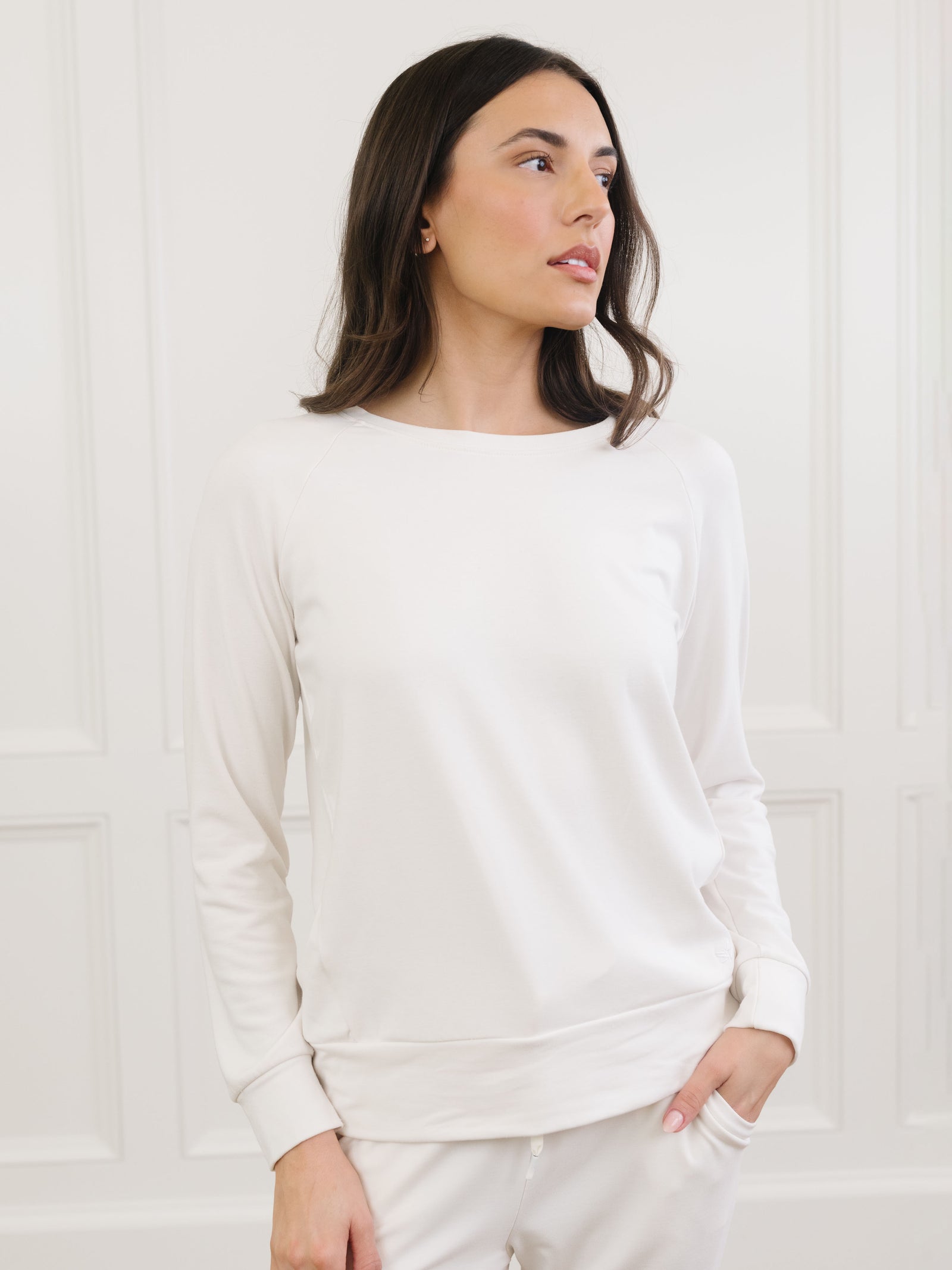 A person with long dark hair is wearing the Women's Ultra-Soft Bamboo Wide Leg Pull On Pant & Pullover Crew Set by Cozy Earth, standing against a white paneled wall. They look to the side with one hand in their pocket. 