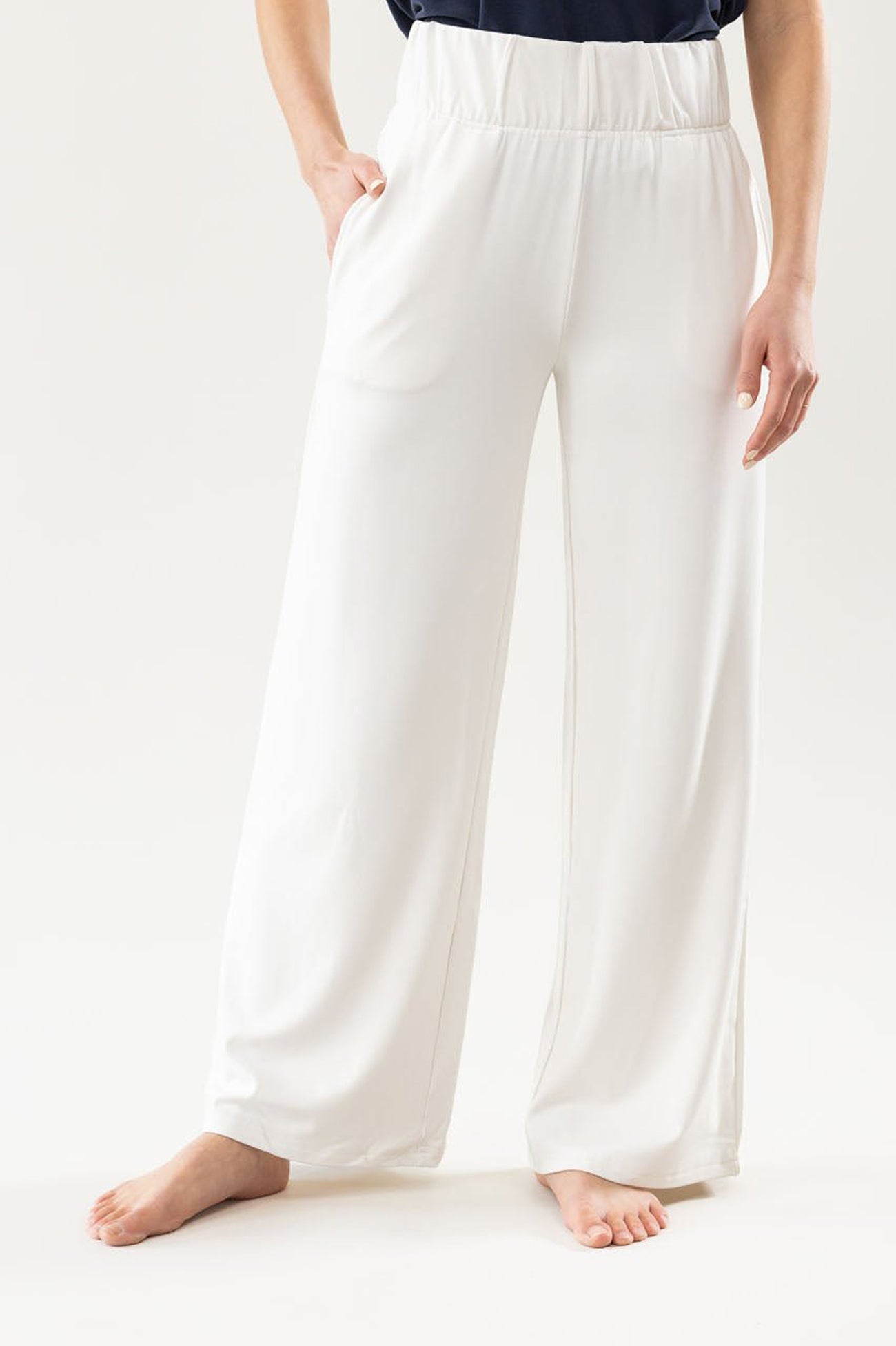 A person wearing loose white pants from Cozy Earth's Women's Ultra-Soft Bamboo Wide Leg Pull On Pant & Hoodie Set stands barefoot with a hand in one pocket against a plain light background, focusing on the pants and lower half of the body. 