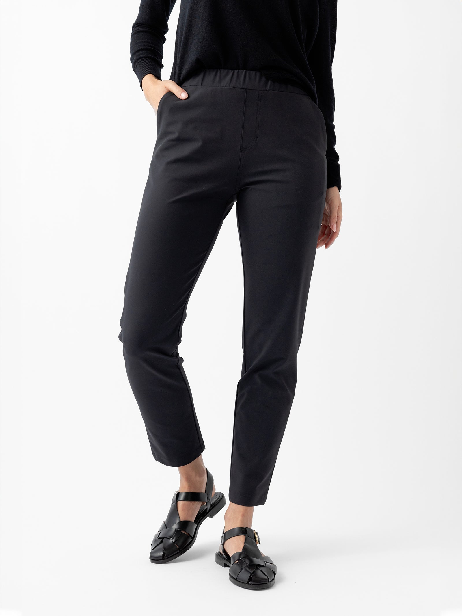 A person wearing Cozy Earth's Women's Always Cropped Pant in black and a black long-sleeve top is standing with one hand in their pocket. The person is also wearing black, open-back, closed-toe loafers. The background is plain white. 