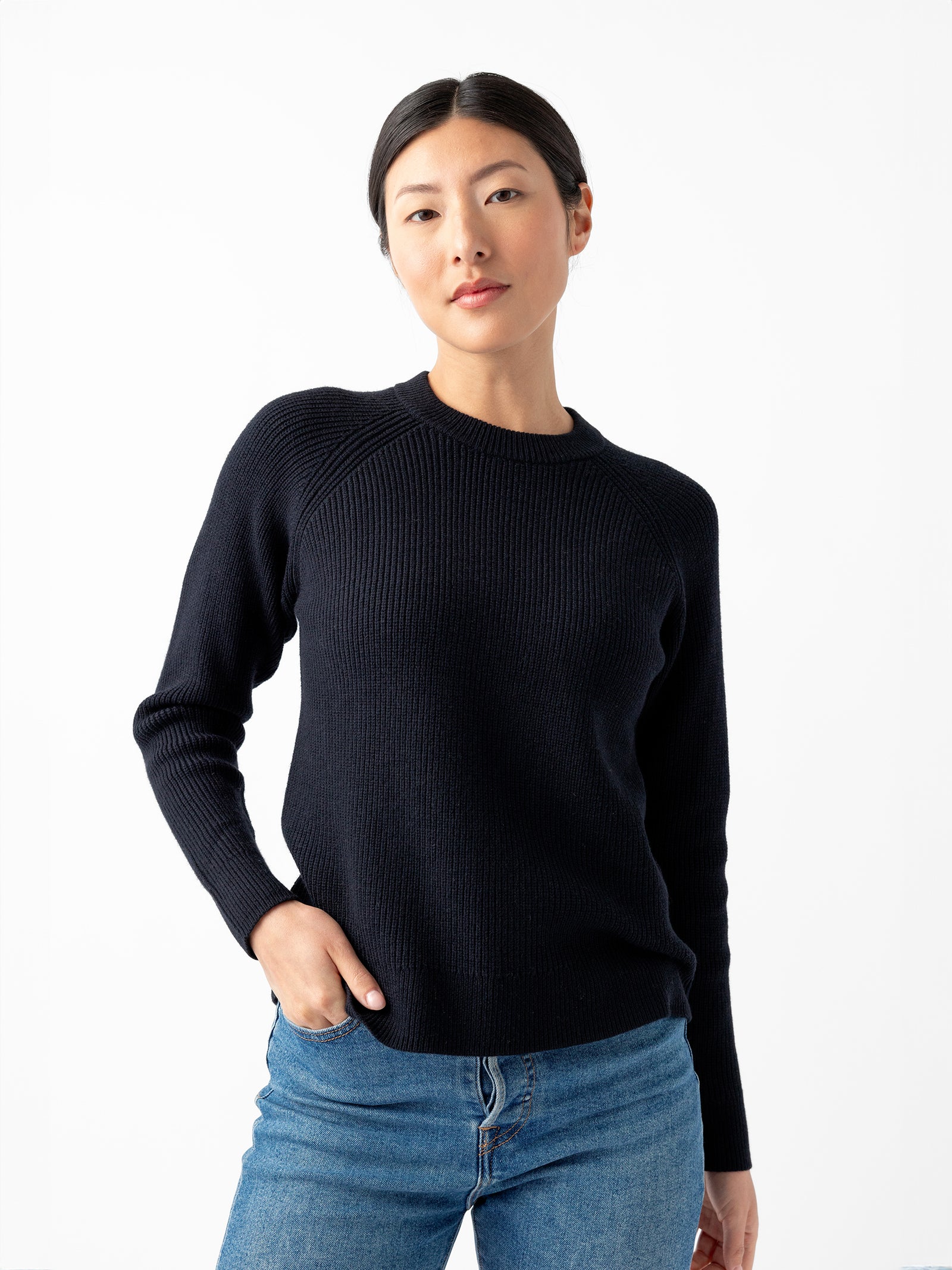 A woman with straight, dark hair is standing against a plain white background. She is wearing Cozy Earth's Women's Classic Crewneck, a knitted long-sleeve dark sweater, paired with blue jeans. Her left hand is in her pocket, and she maintains a neutral expression with her lips closed. 