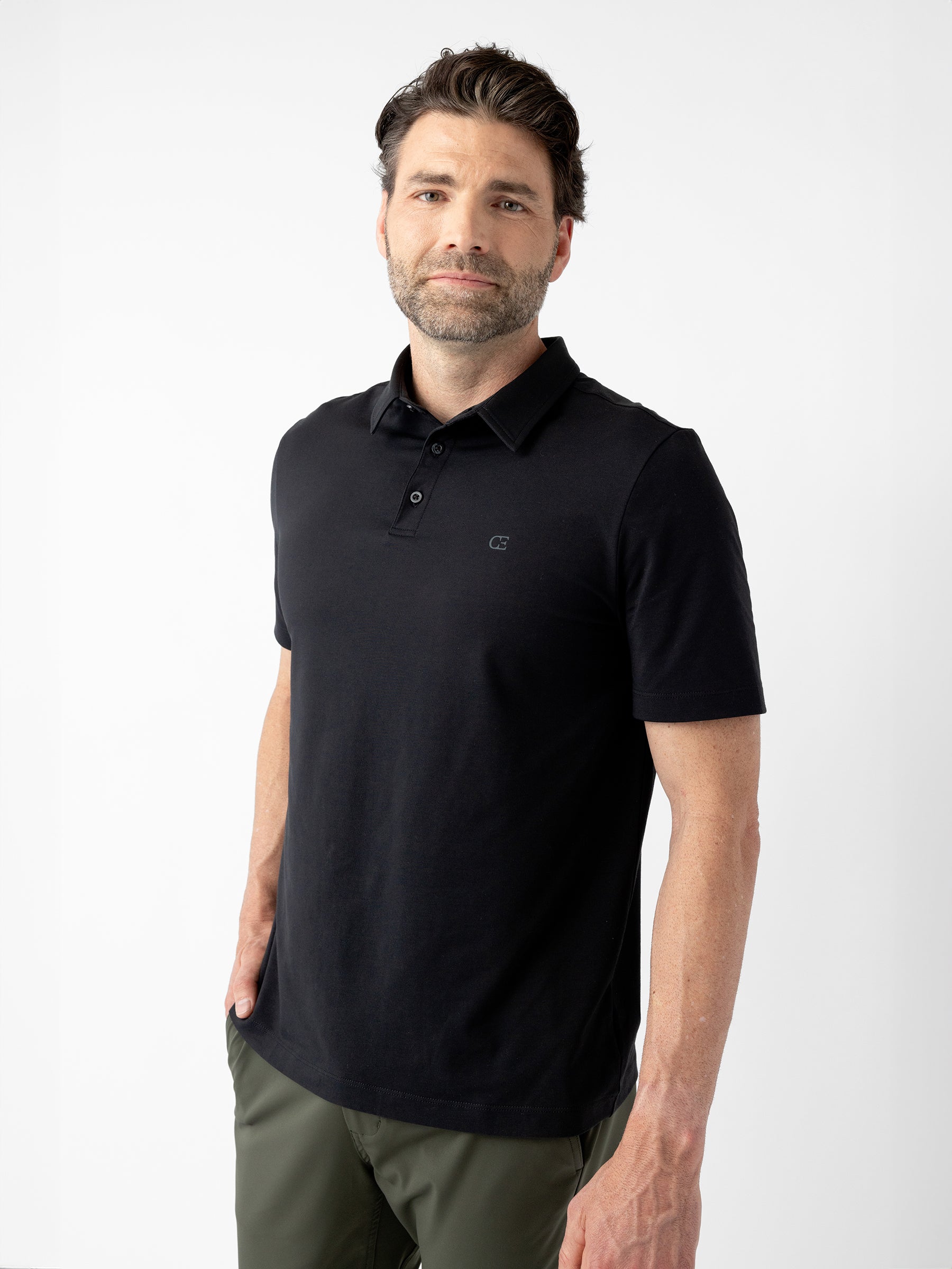 A man with short, dark hair and a beard wears a Men's Everyday Polo by Cozy Earth in black paired with dark green pants. He stands against a plain white background with his right hand in his pocket and a neutral expression on his face. |Color:Jet Black