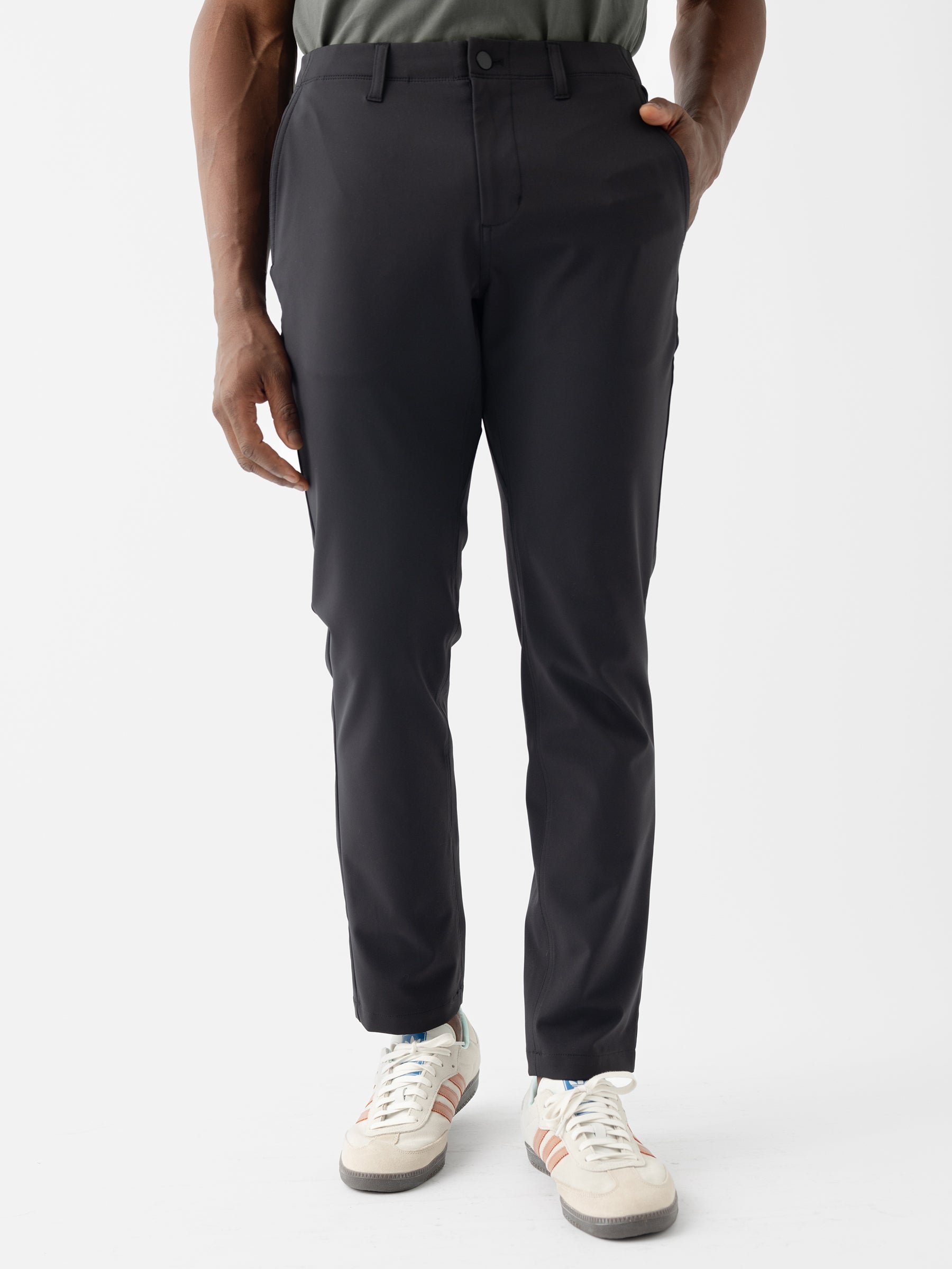 A person is standing against a white background with their right hand in their pocket. They are wearing Cozy Earth's Men's Everywhere Pant 30L in a dark color, a grey shirt, and white sneakers with pastel-colored accents. The pants have a classic fit with a waistband and front zipper. |Color:Jet Black
